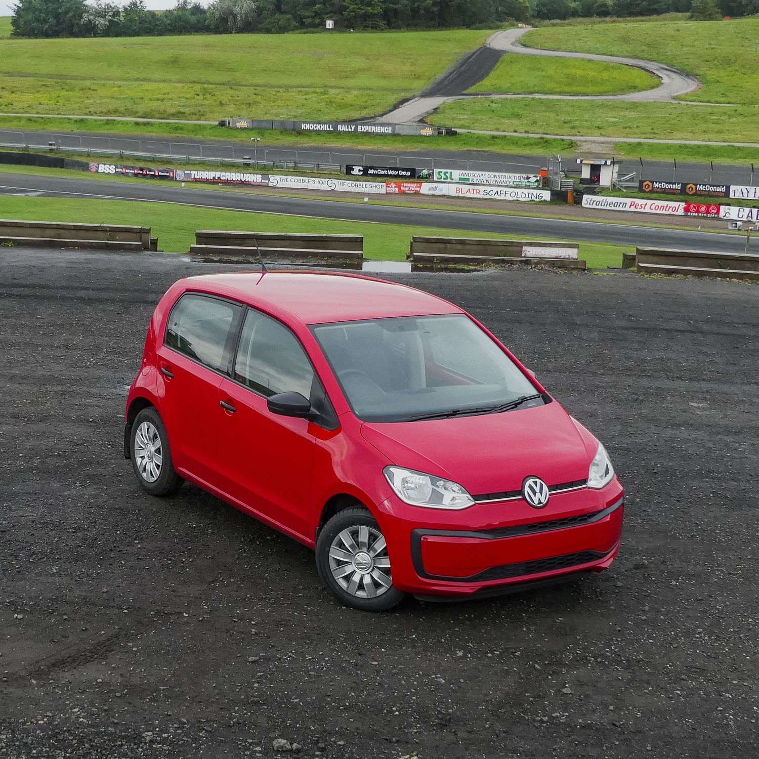 A bright red Volkswagen Up compact car parked on a race track with winding course and green fields in the background.