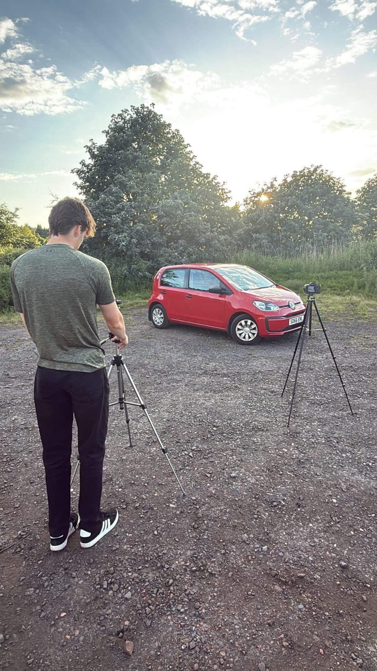 Photographer with tripod capturing a red Volkswagen hatchback parked on gravel at sunset with lush trees in background.