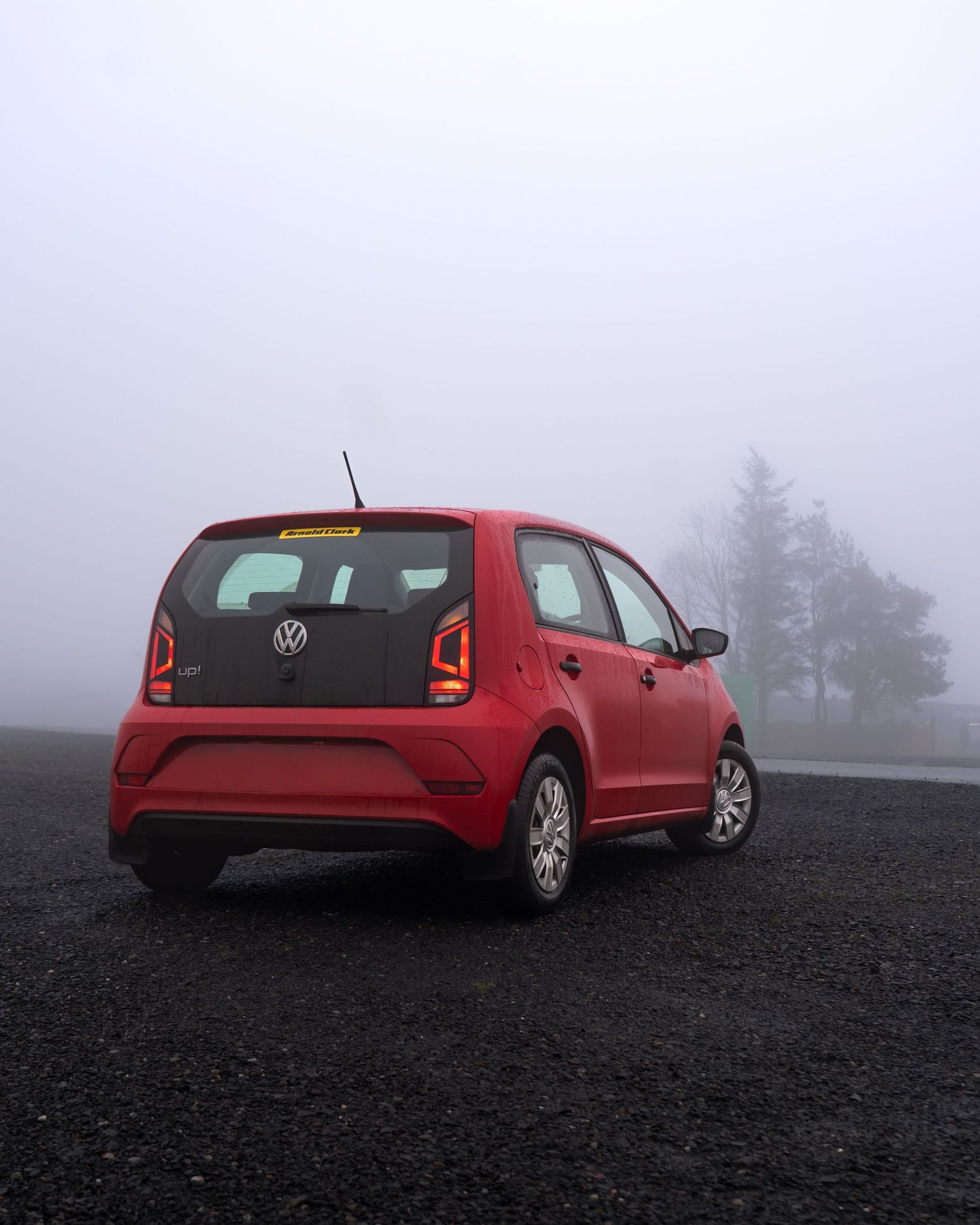 A red car sits on black volcanic soil in foggy conditions with pine trees visible in the misty background.