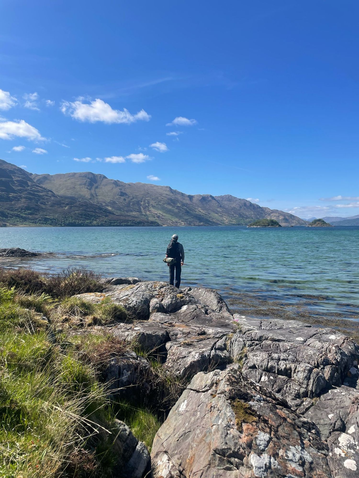 Person standing on rocky shoreline gazing at a serene Scottish loch with mountains and blue sky backdrop.