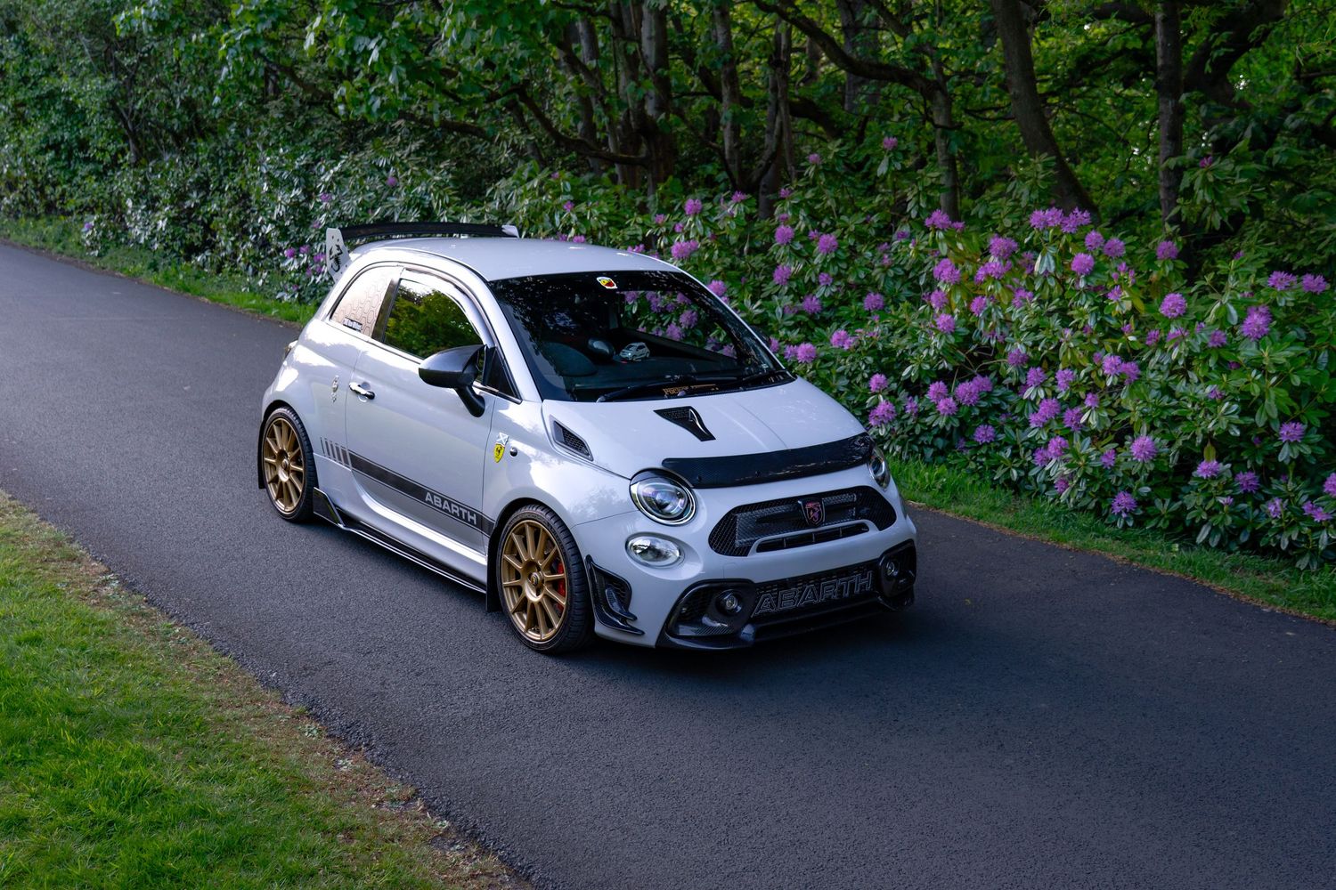 Modified Abarth 595 in silver with gold alloy wheels on a winding road surrounded by lush green hedges and purple flowers.