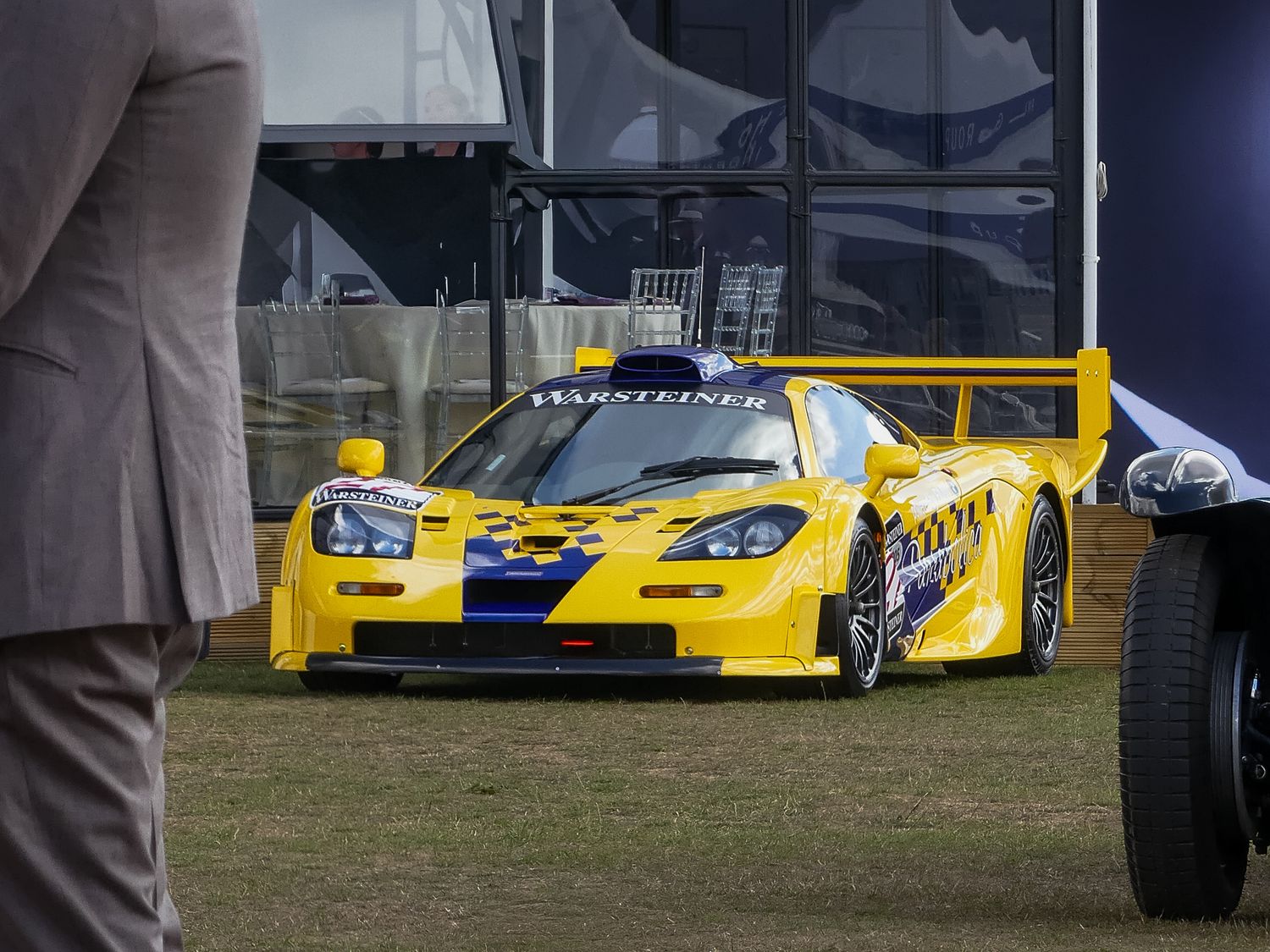 Yellow racing car parked on grass field with other vehicles visible in background during motorsport event.
