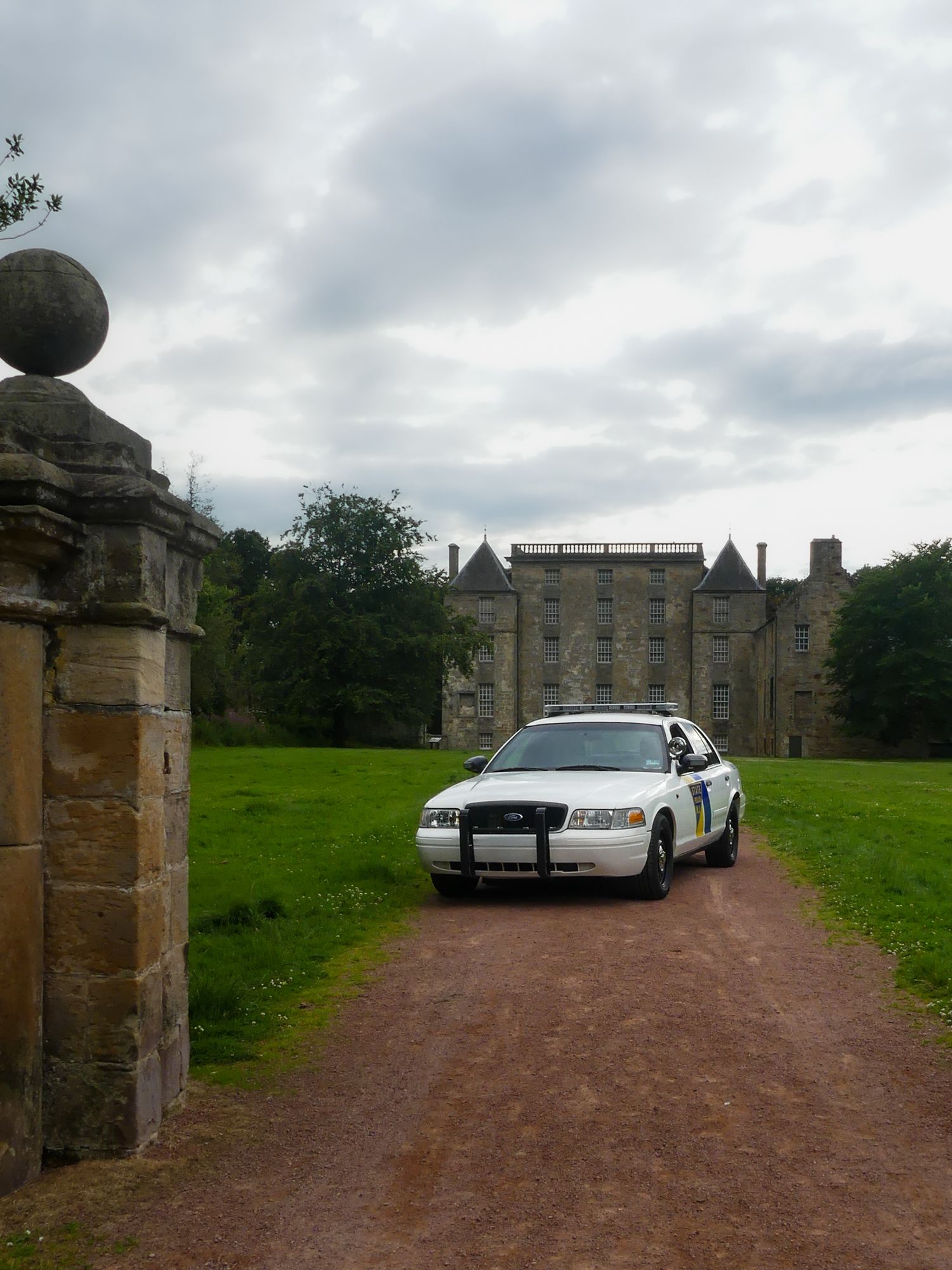 A police car parked on a gravel driveway in front of a historic stone mansion with cloudy skies overhead.