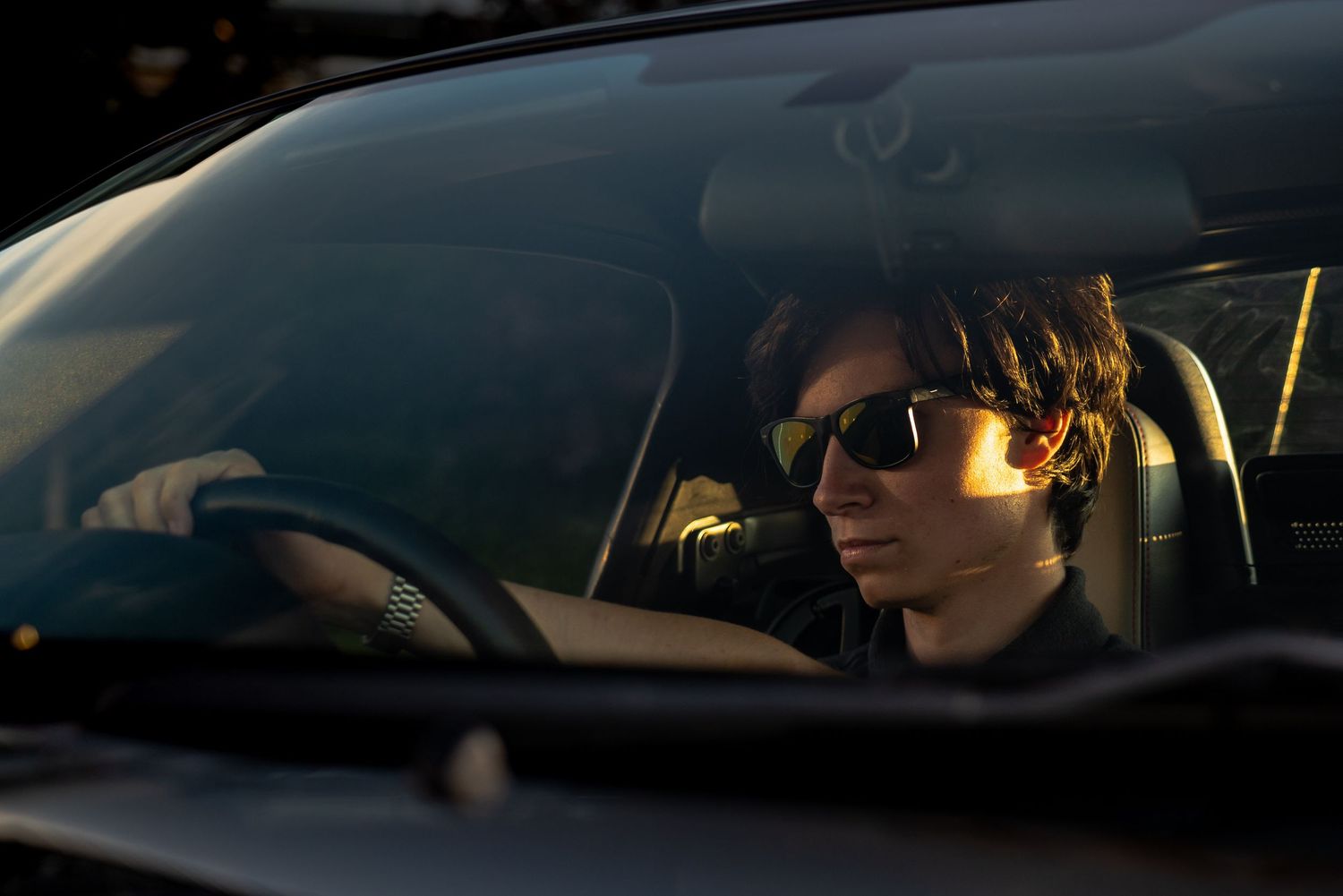 Young man wearing sunglasses driving a car, illuminated by warm sunlight through the windshield.