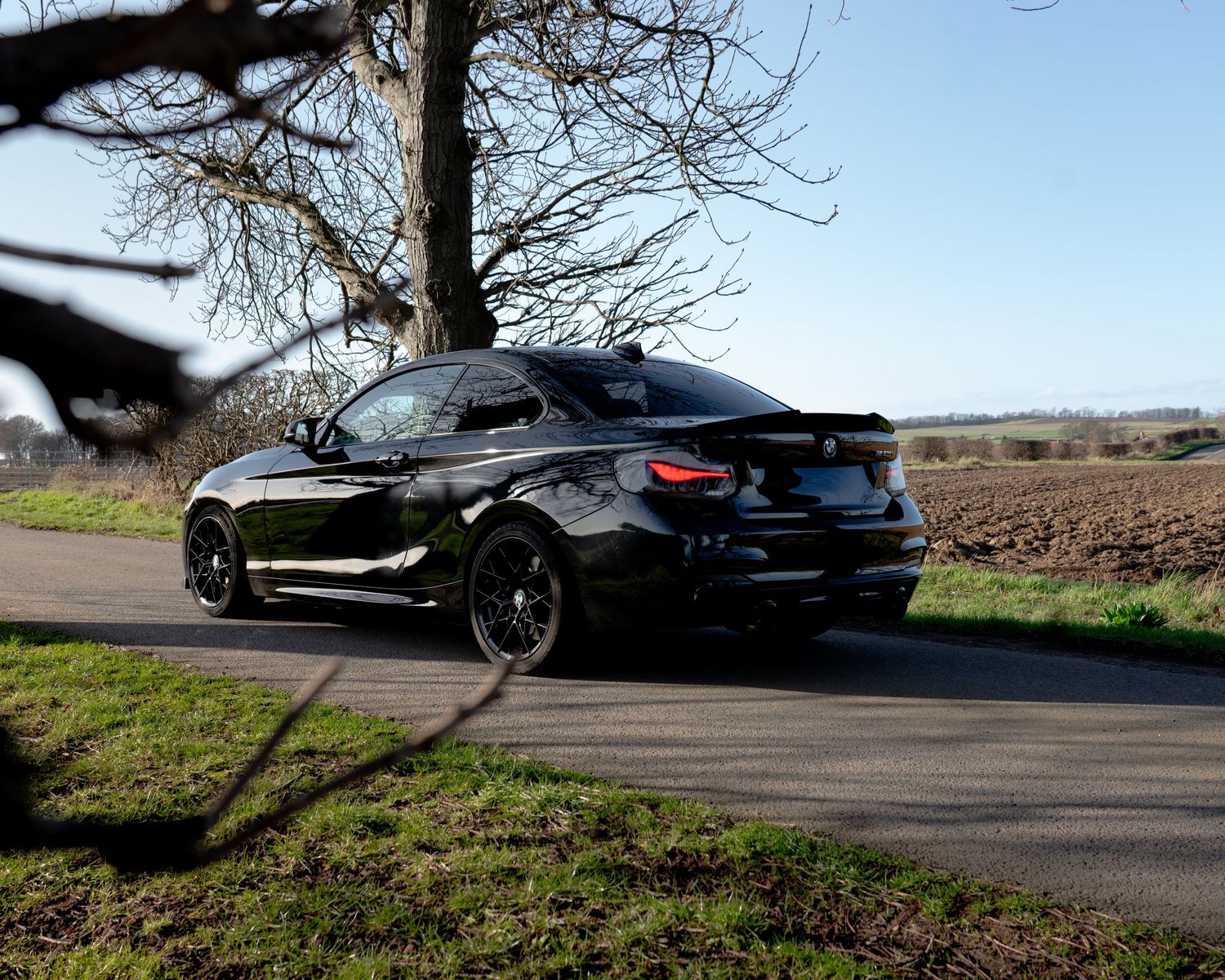 Black BMW M4 with dark wheels parked on a country road with fields and trees in the background.