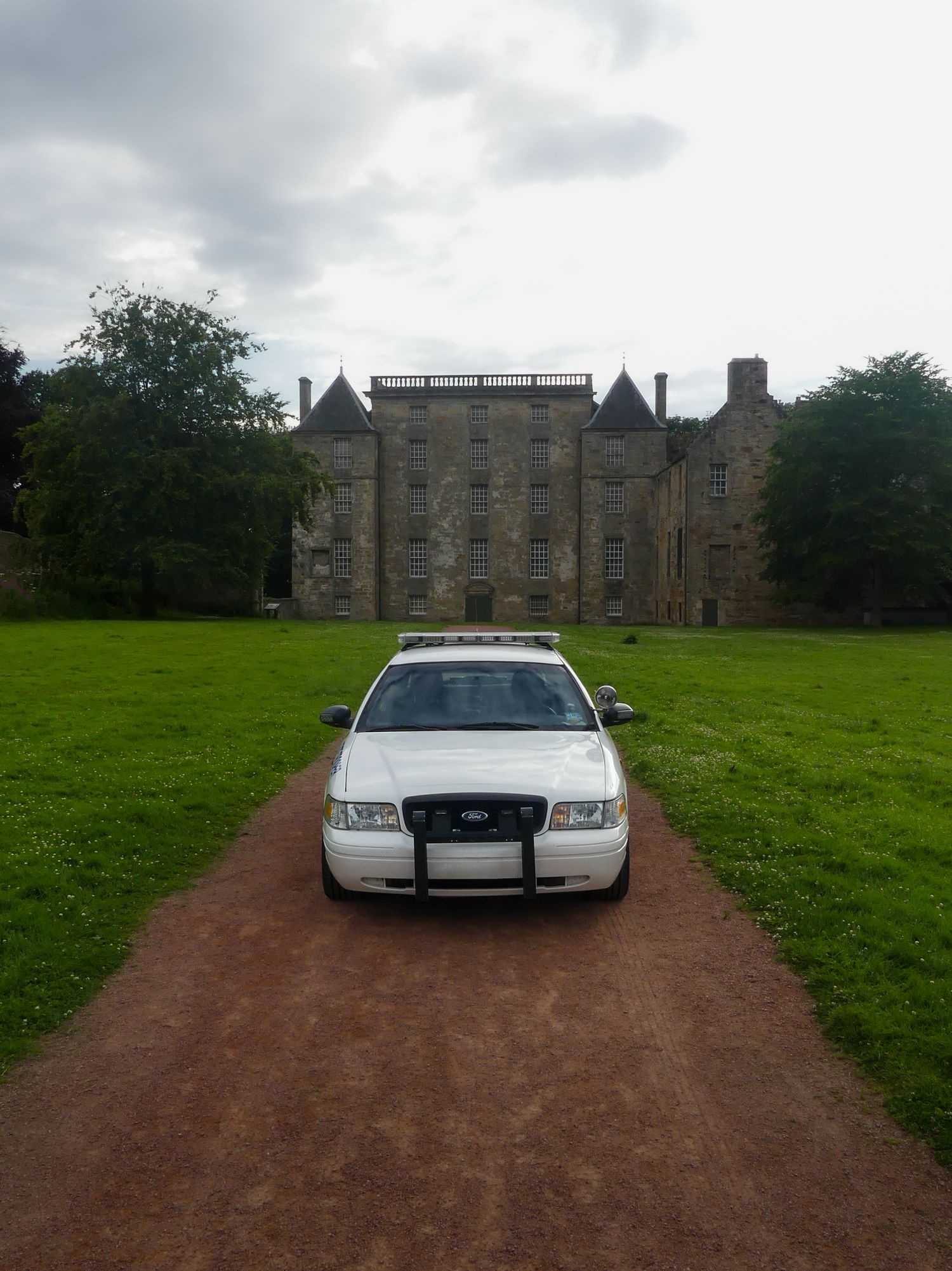 White police cruiser centered on a dirt path leading to a gothic-style stone castle under cloudy skies.