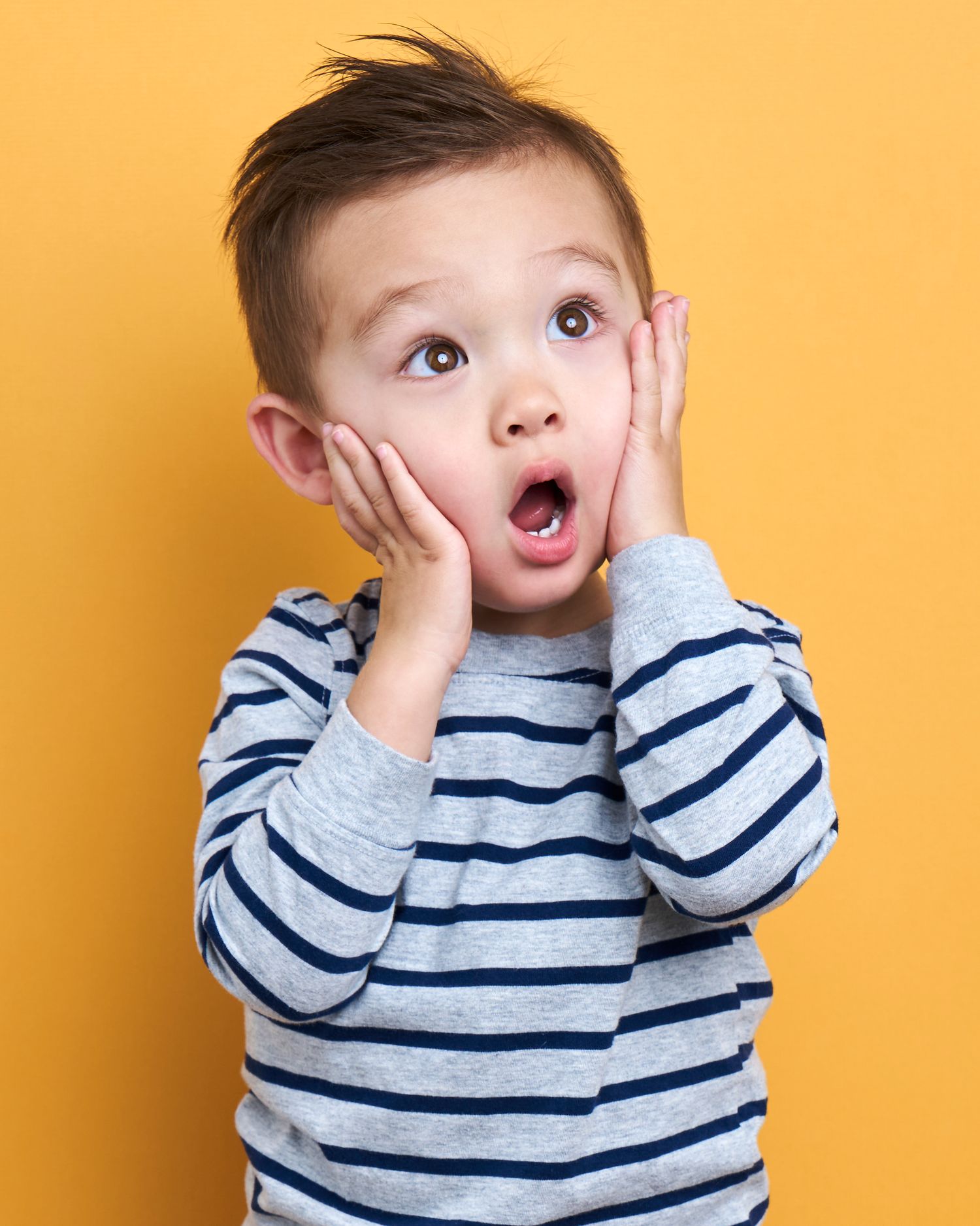 Young child in striped shirt making playful expressions against orange background.