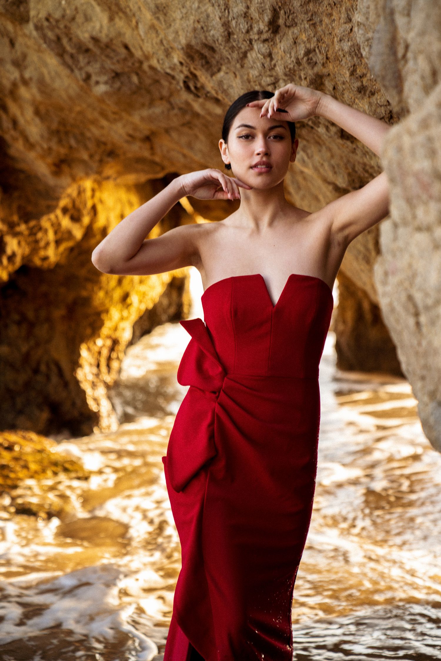 Model in red evening gown poses dramatically in coastal cave setting.