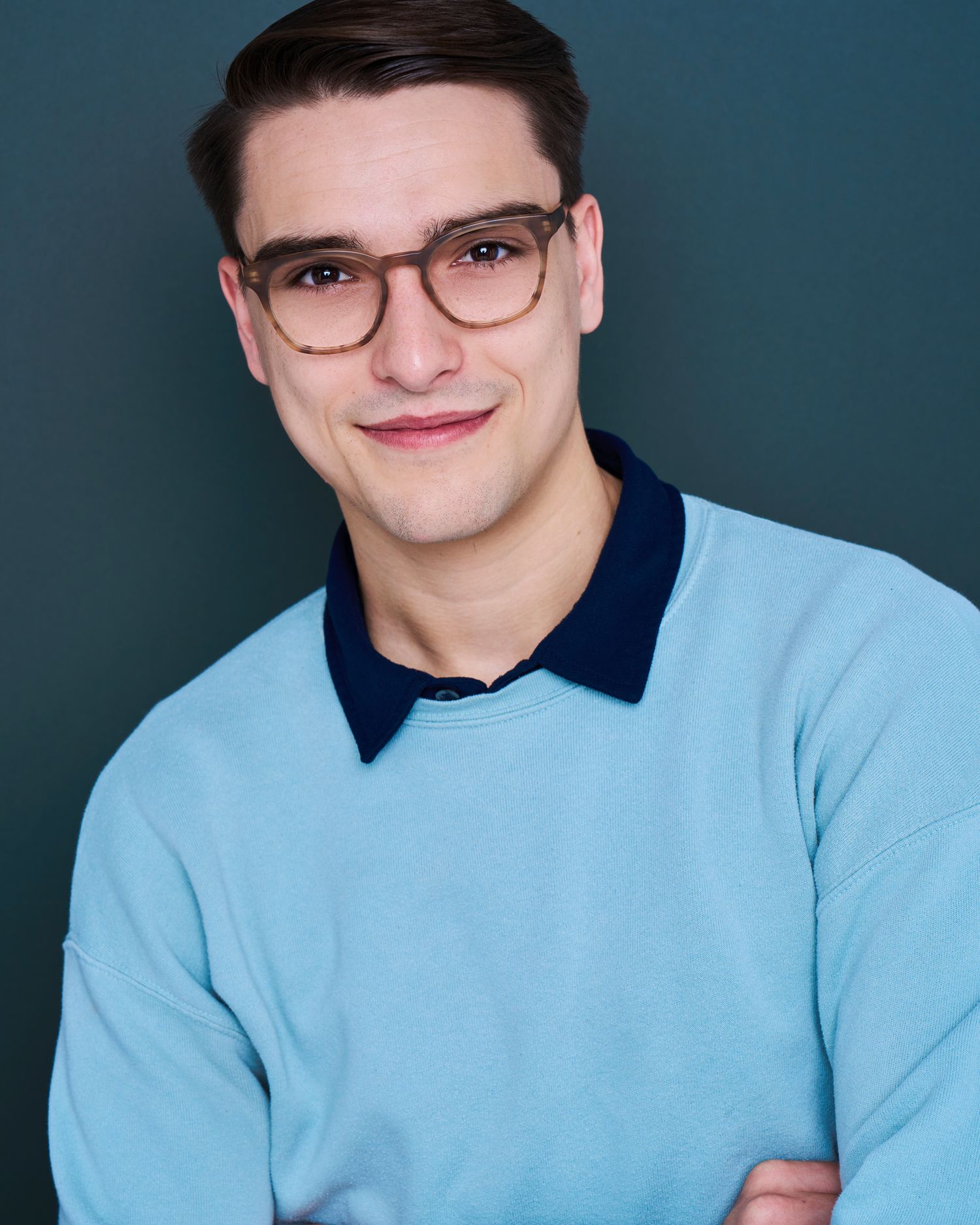Studio portraits wearing light blue sweater with navy collar against dark blue background.