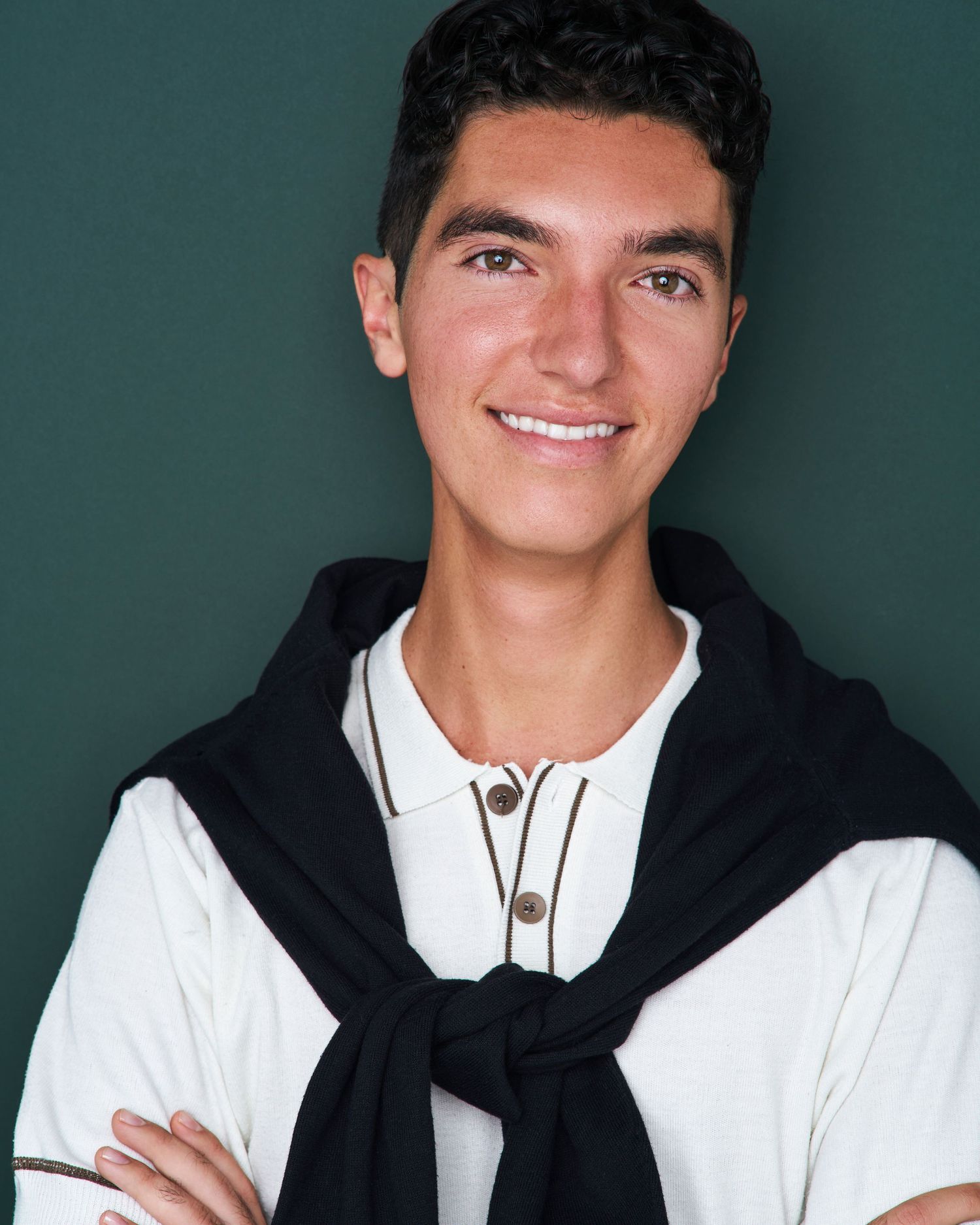 Professional headshot of a smiling person in a white shirt with black sweater tied around shoulders against green background.