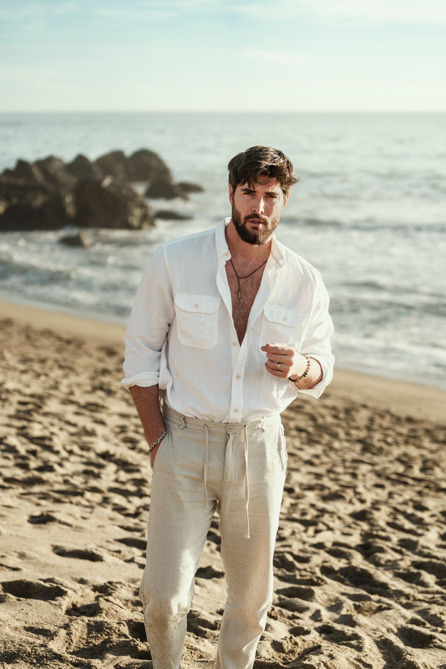 Person wearing all-white outfit walking along rocky beach coastline at sunset.