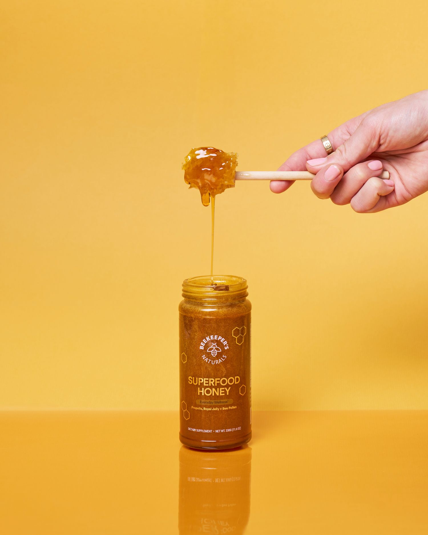 Hand drizzling golden honey from a glass jar against a vibrant yellow background in a minimalist product shot.