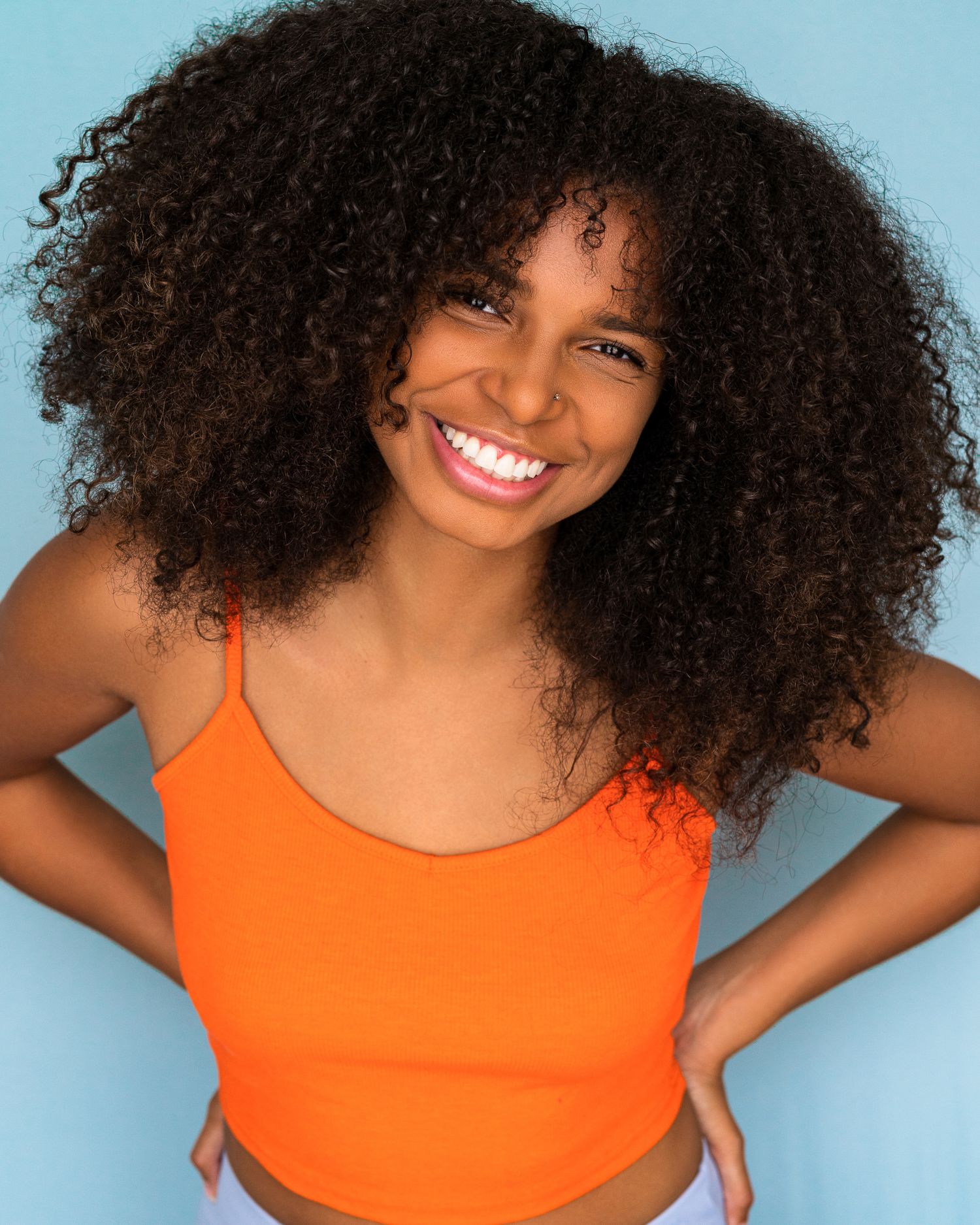Woman with natural curly hair smiles brightly wearing an orange tank top against blue background.