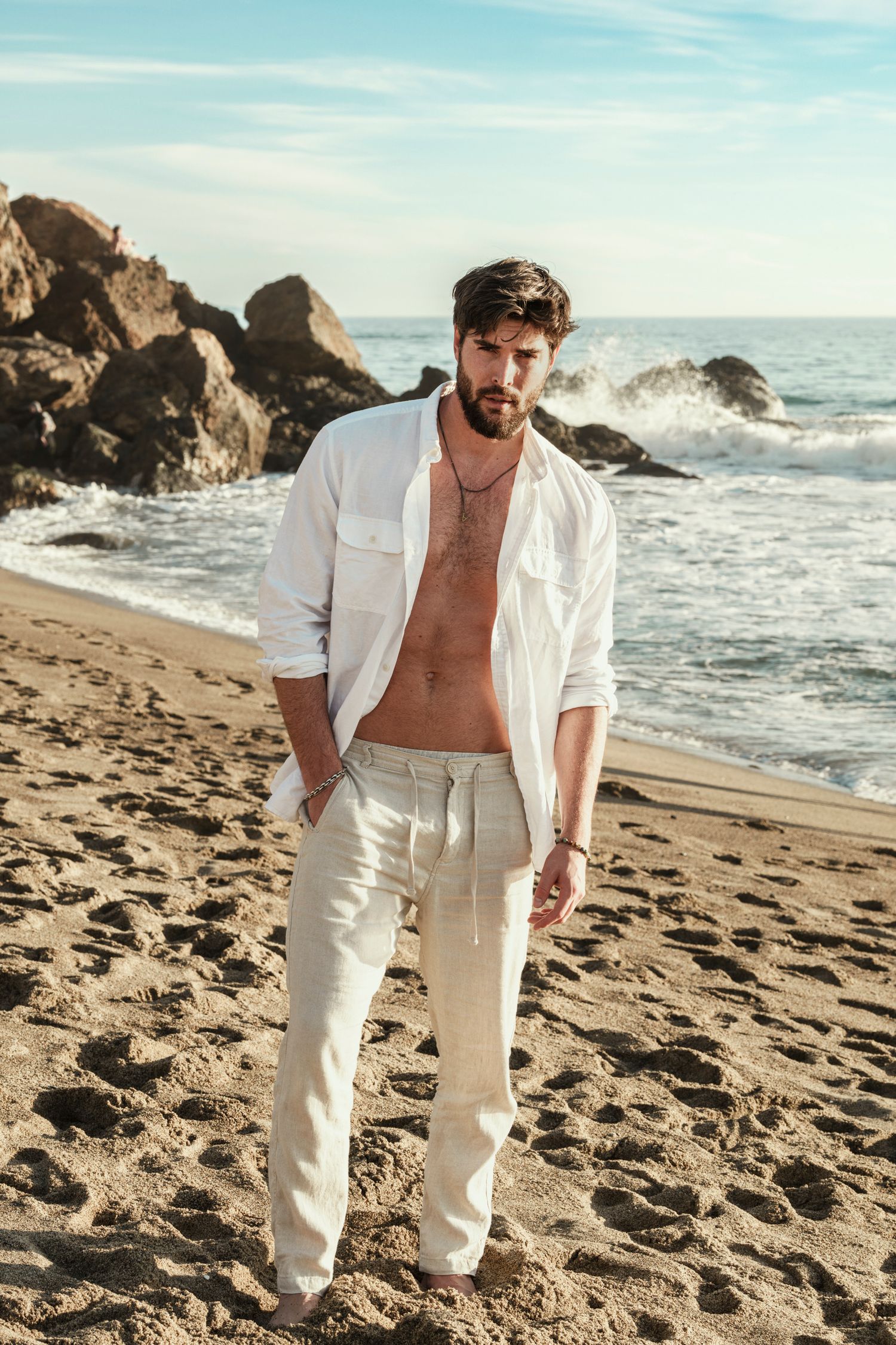 Model in all-white outfit poses on sandy beach with rocky coastline and ocean waves.