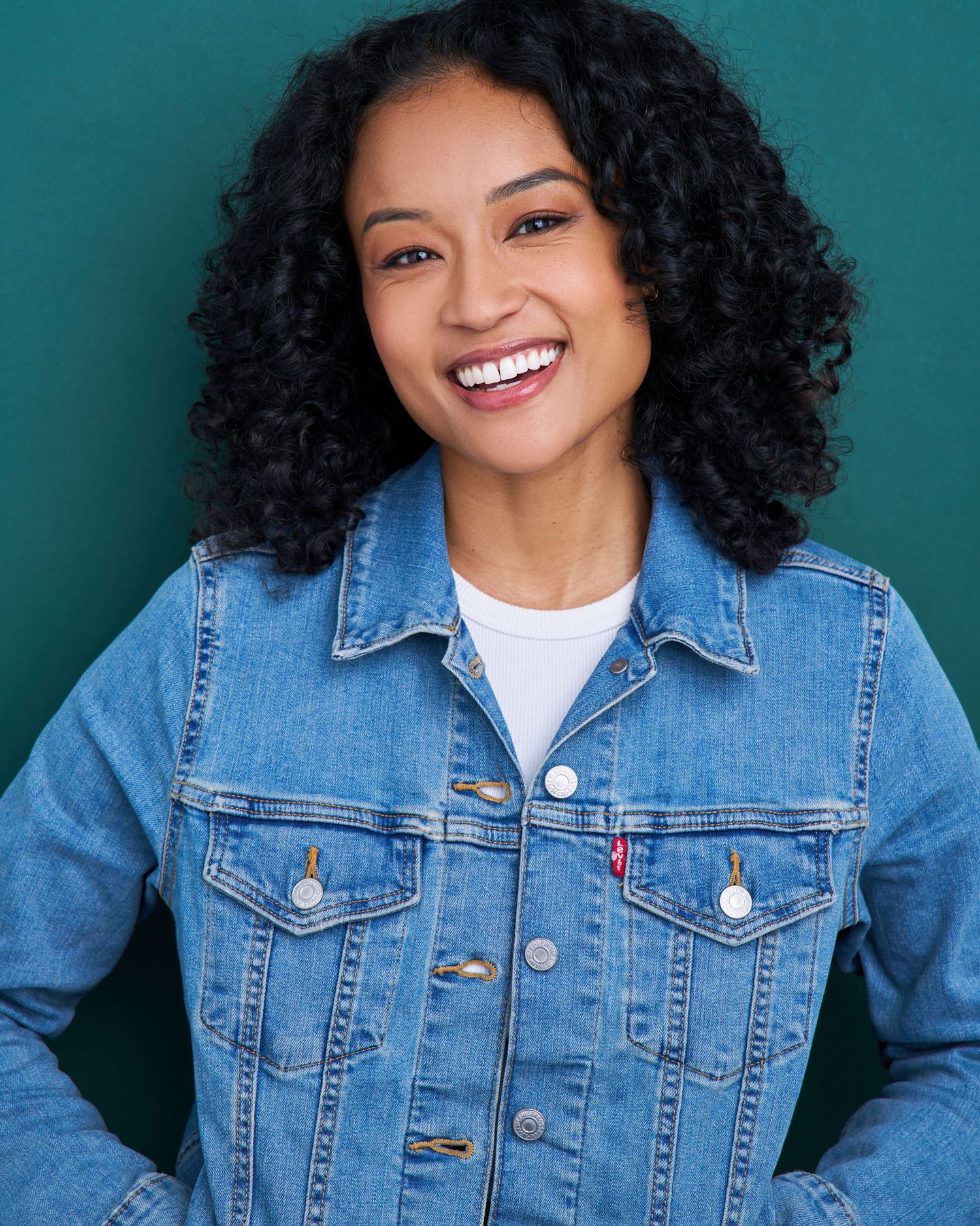 Casual portrait in denim jacket with natural curly hair against teal background.