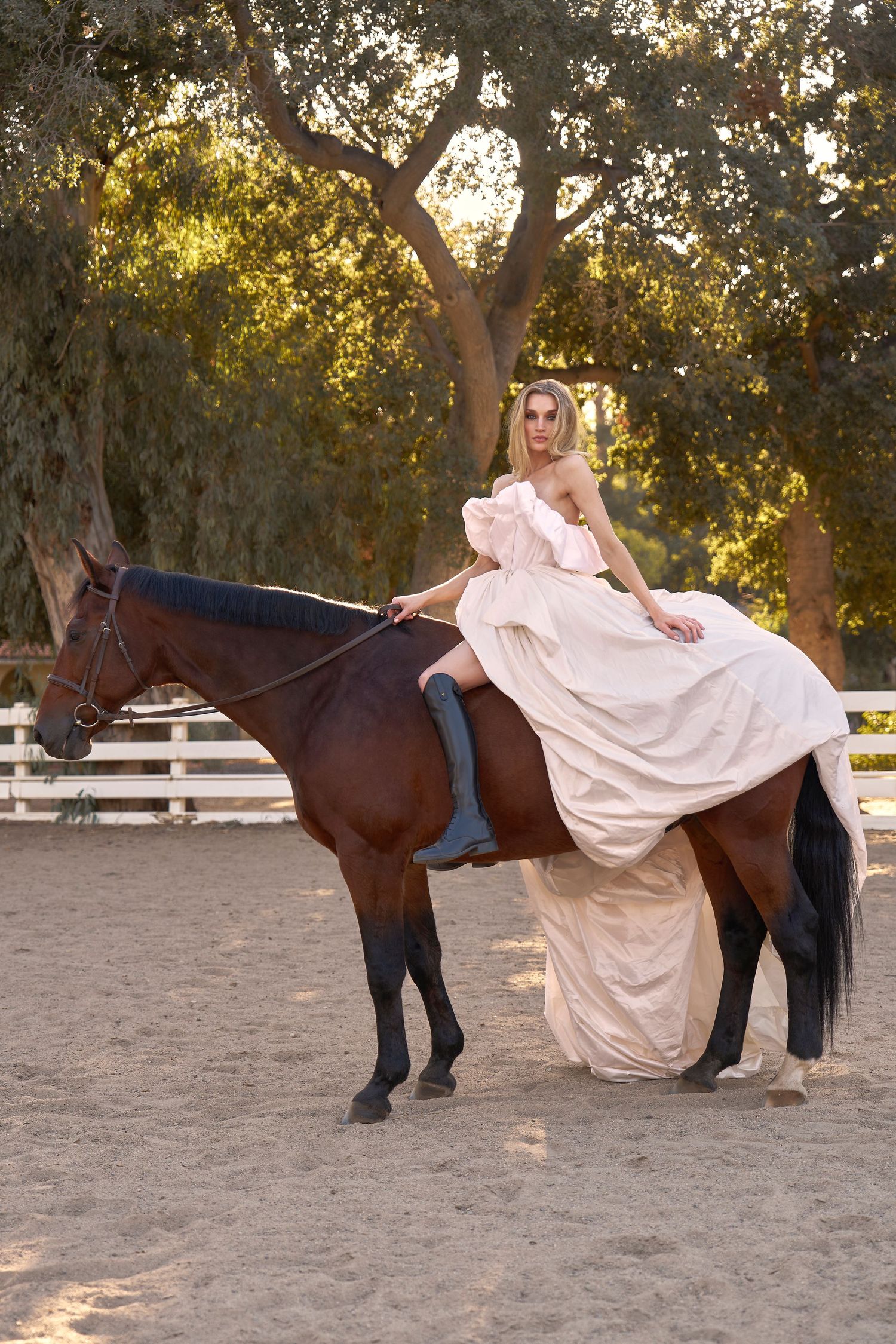 Ethereal photoshoot with flowing white dress and brown horse in outdoor arena at sunset.