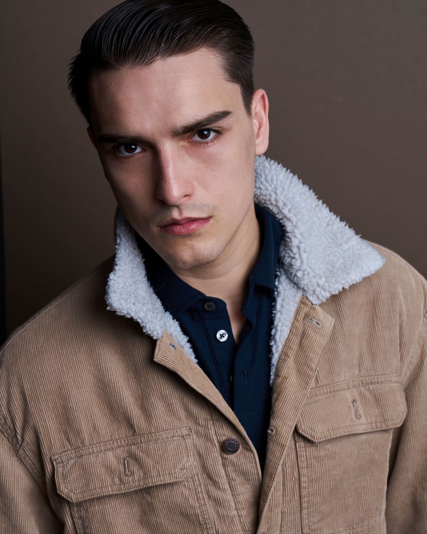 Fashion portraits in tan jacket with sherpa collar and navy shirt against brown background.