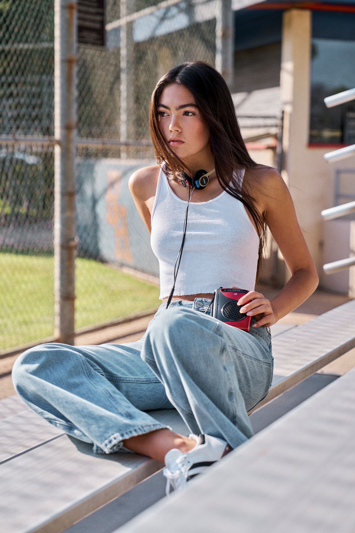 Casual street style with white tank top and relaxed denim by chain link fence.