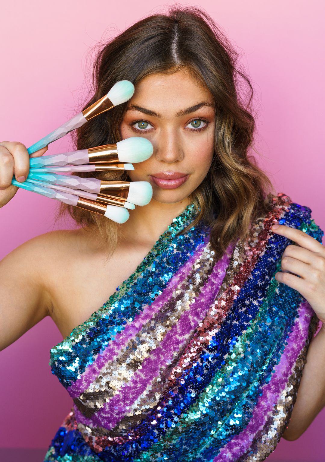Fashion model in sequined dress holding makeup brushes against pink backdrop.