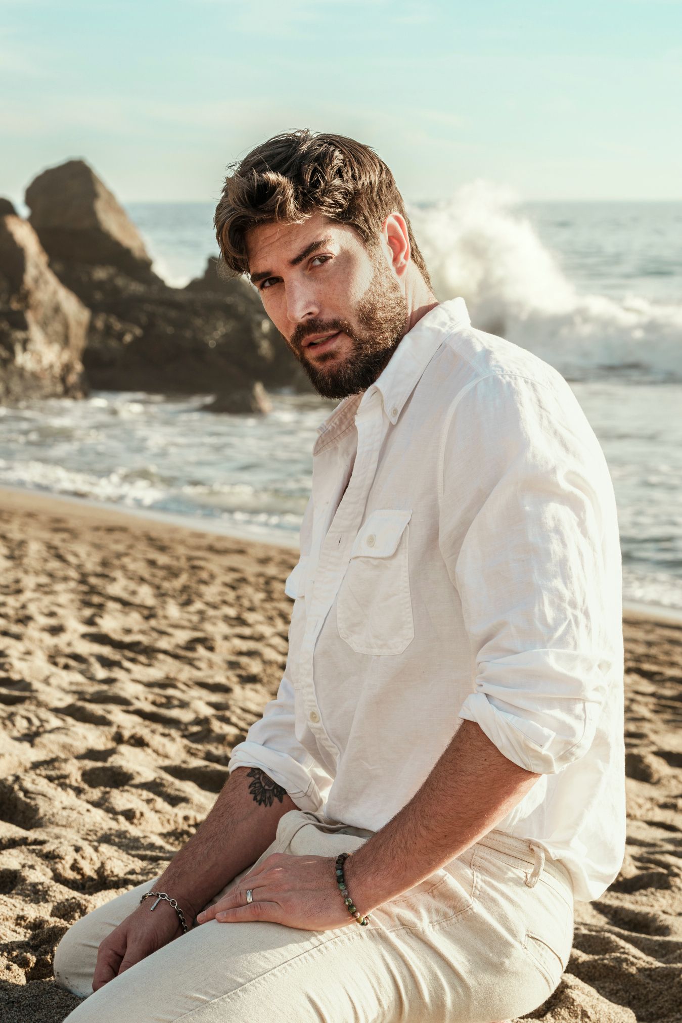 Person in white shirt and pants sitting on beach with crashing waves and rocks in background.