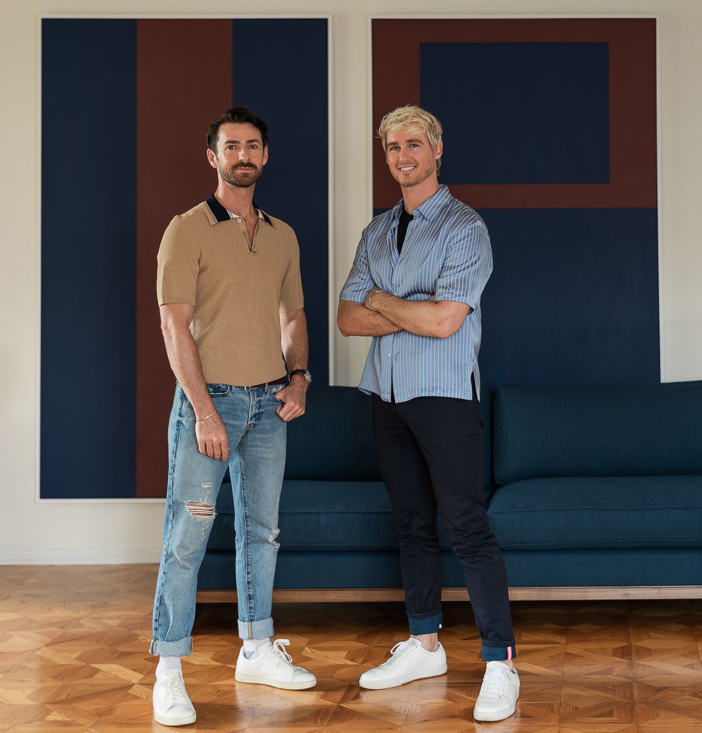Two people standing casually next to a dark sofa in a minimalist room with wooden floors.