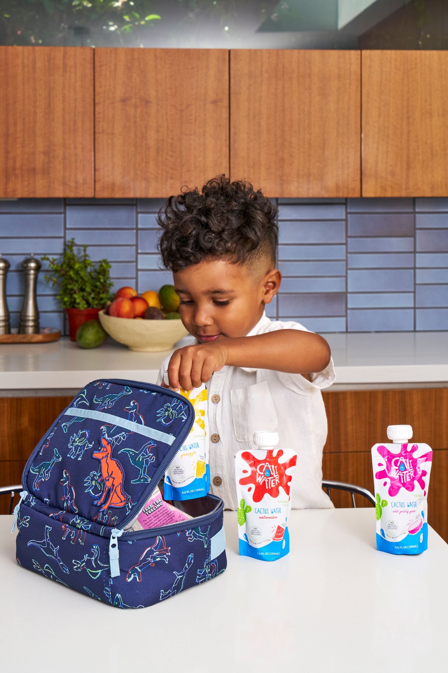 Child packing colorful snack pouches into blue dinosaur-patterned lunch bag in modern kitchen.