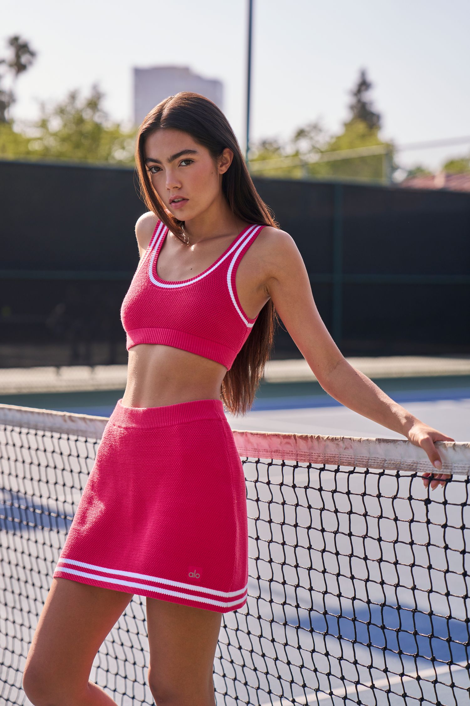 Bright pink athletic two-piece outfit shown against tennis court net and fence.