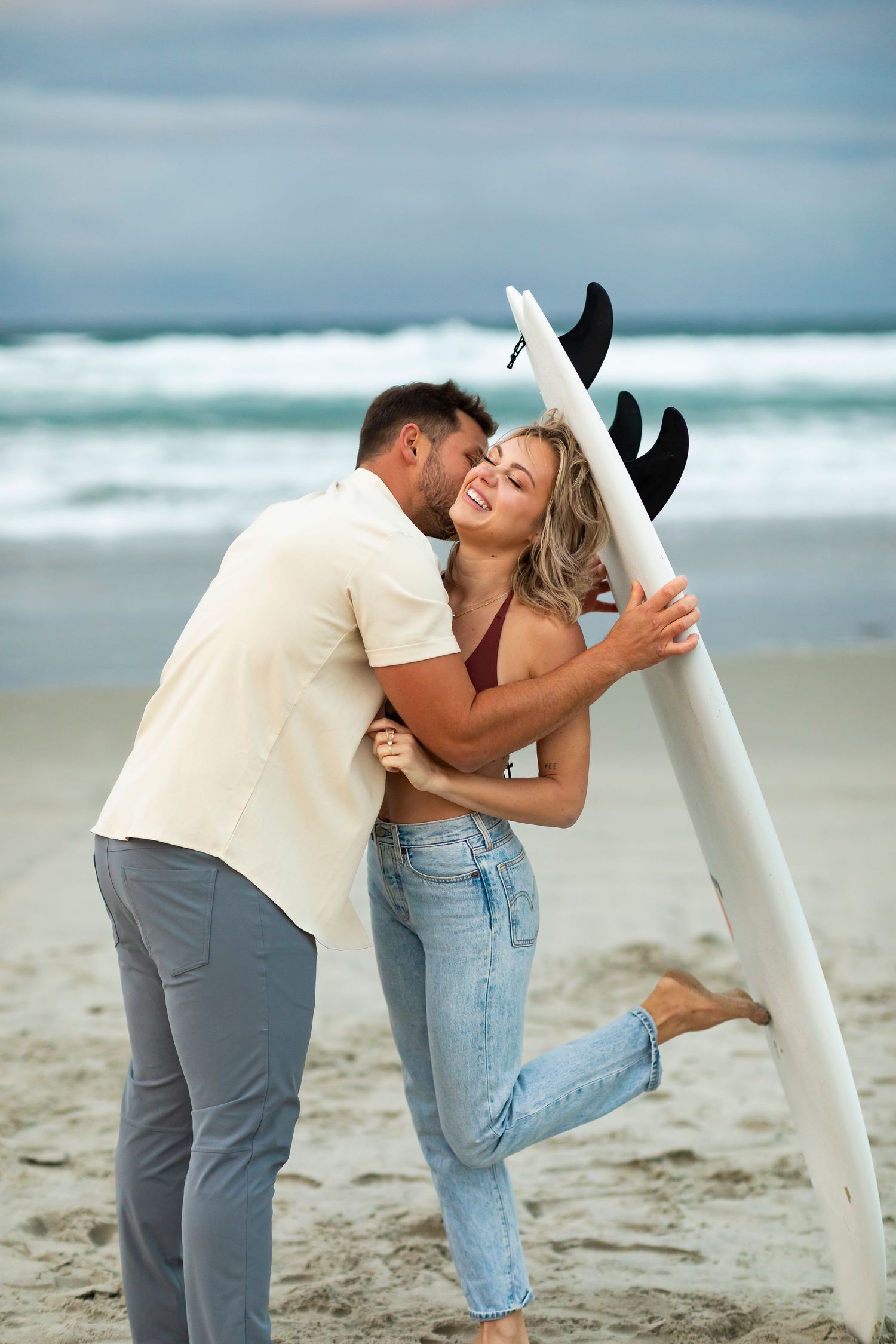 destination photoshoot of beach engagement new zealand couple carries surfboard