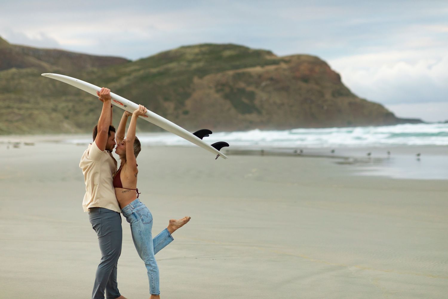 destination photoshoot of beach engagement new zealand couple carries surfboard