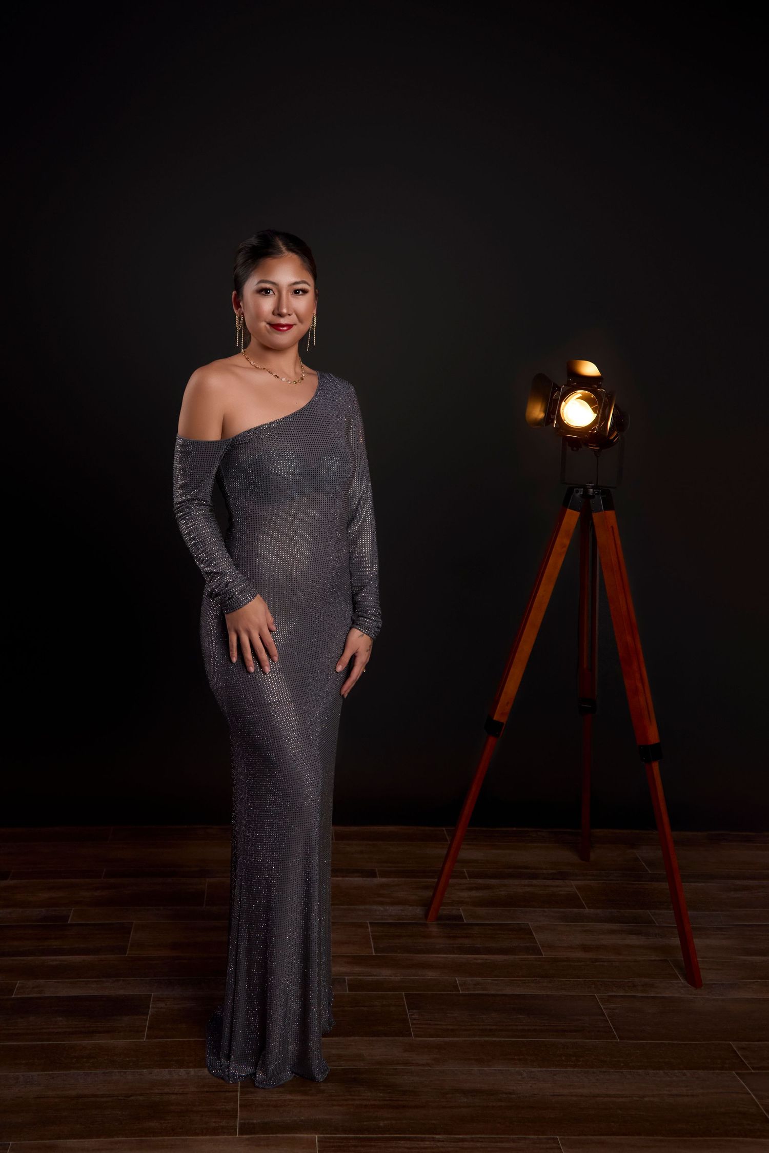Elegant one-shoulder gray evening gown shown in dramatic studio setting with vintage tripod light fixture on hardwood floor.