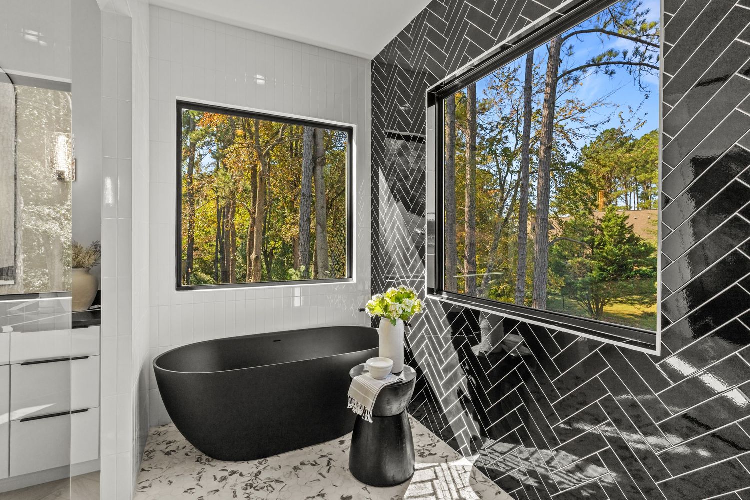 Modern bathroom with black freestanding tub, herringbone tile, and large windows overlooking autumn forest views.