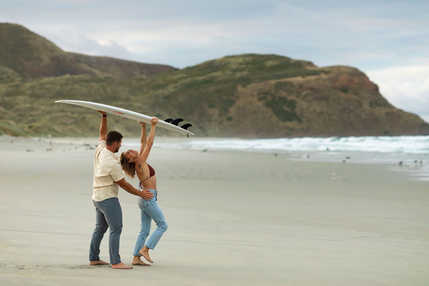 destination photoshoot of beach engagement new zealand couple carries surfboard