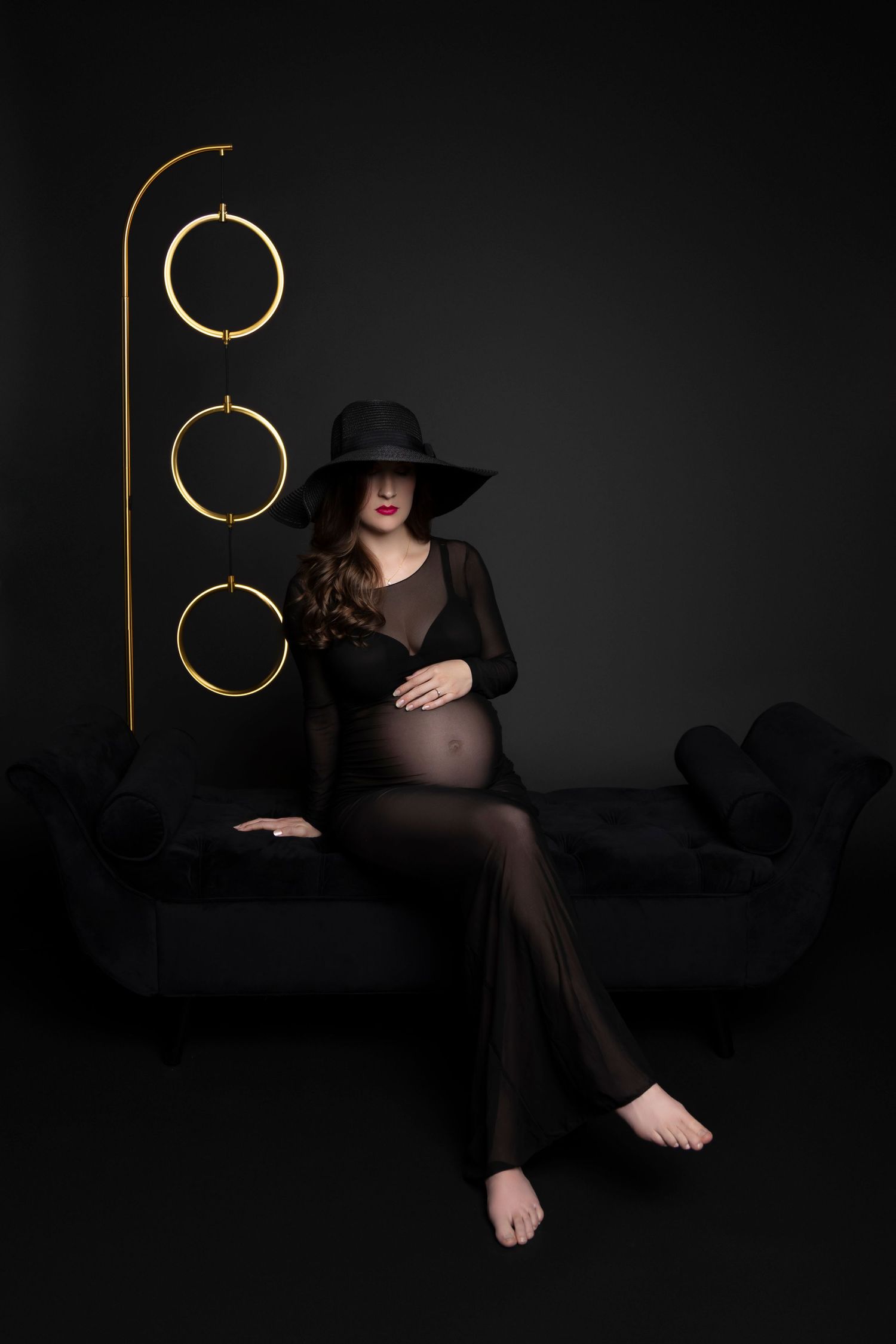 Artistic maternity photo with black dress and hat against dark background with decorative golden circles on stand.