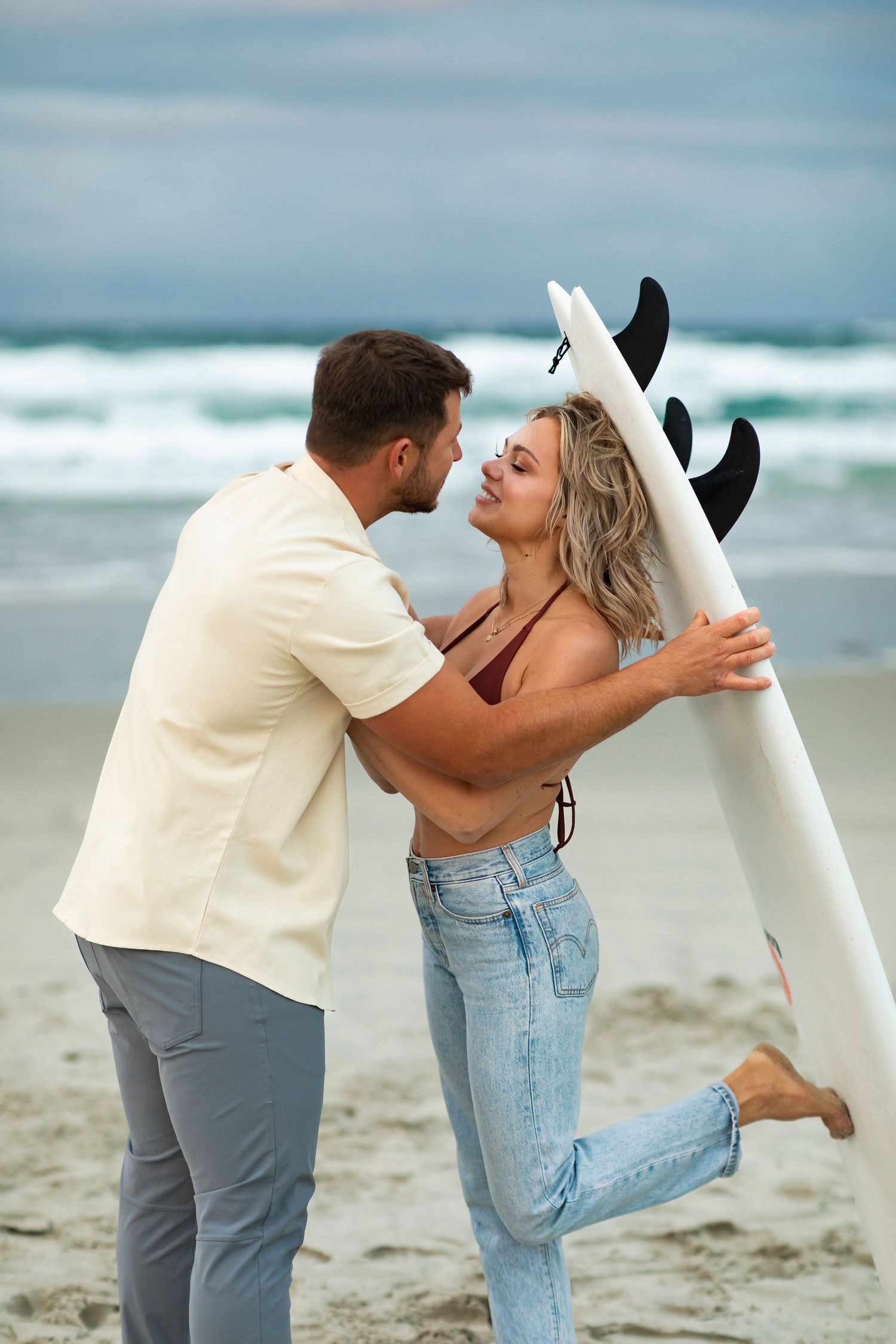destination photoshoot of beach engagement new zealand couple carries surfboard