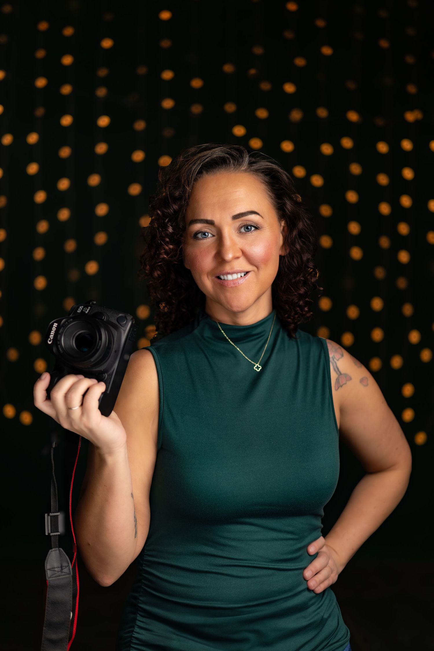 Professional photographer in dark green sleeveless top poses with camera against bokeh background.