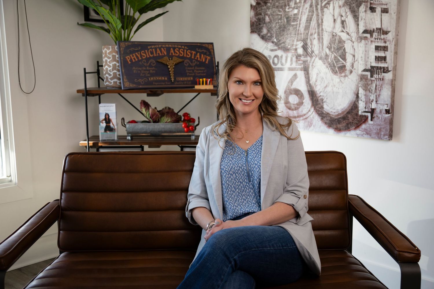 A person sits on a brown leather bench wearing a gray blazer and blue top in an office setting with vintage decor.