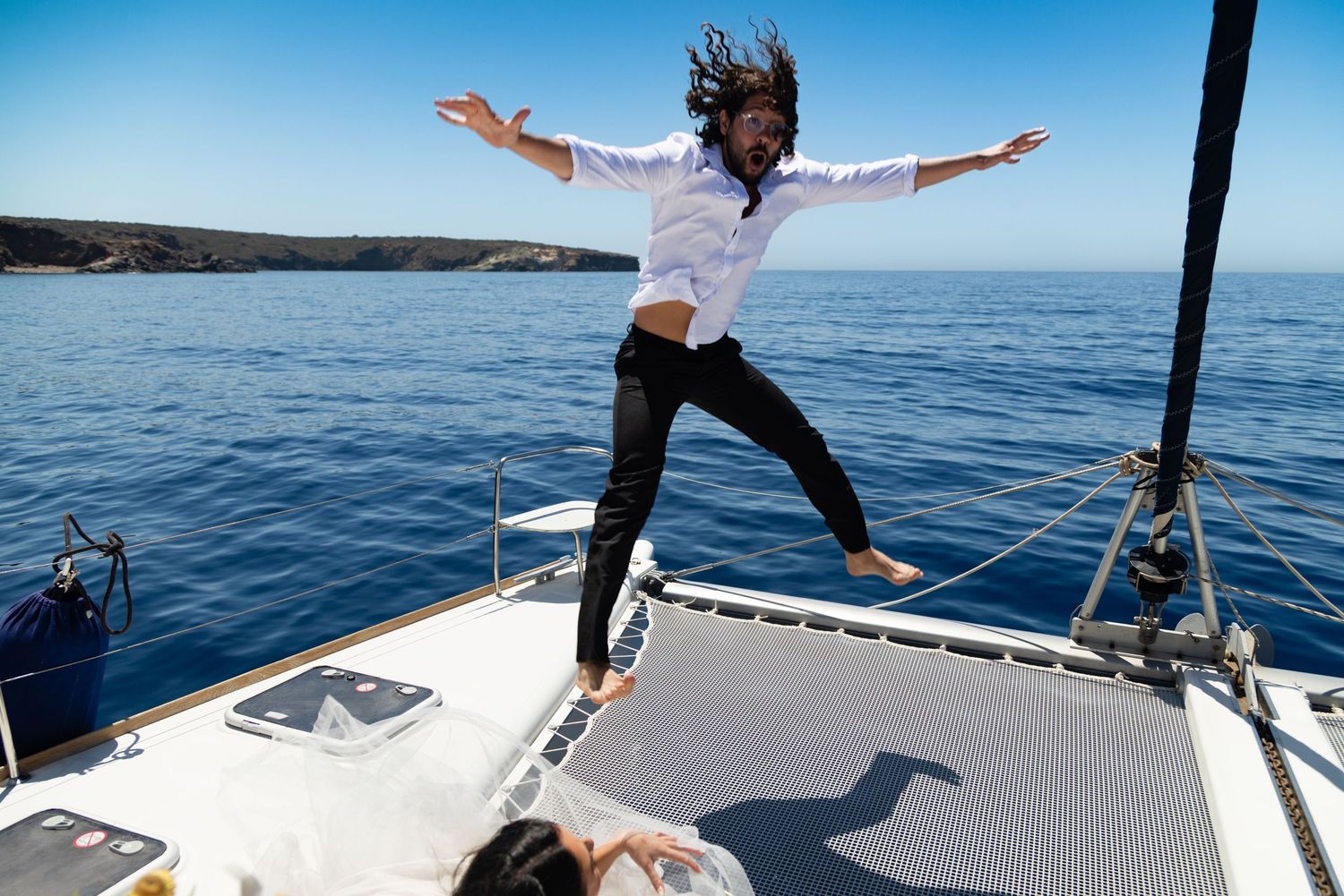 Joyful person jumps with outstretched arms on a catamaran deck sailing across bright blue ocean waters.