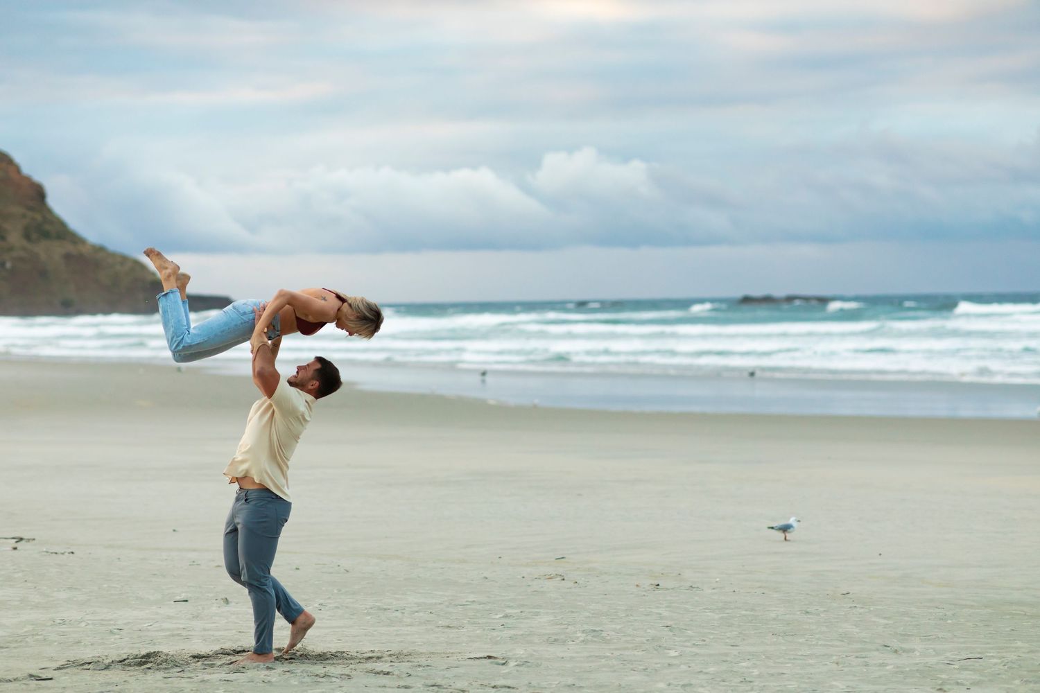 A couple plays and embraces on a scenic beach with waves and cliffs in the background.