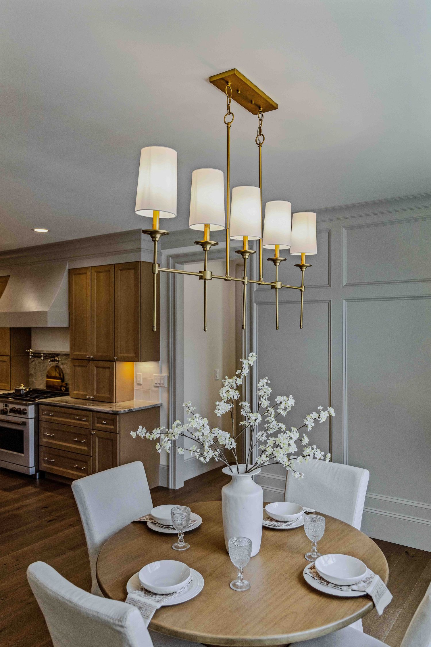 Modern dining area with round wooden table, white chairs, brass chandelier, and elegant white floral centerpiece.
