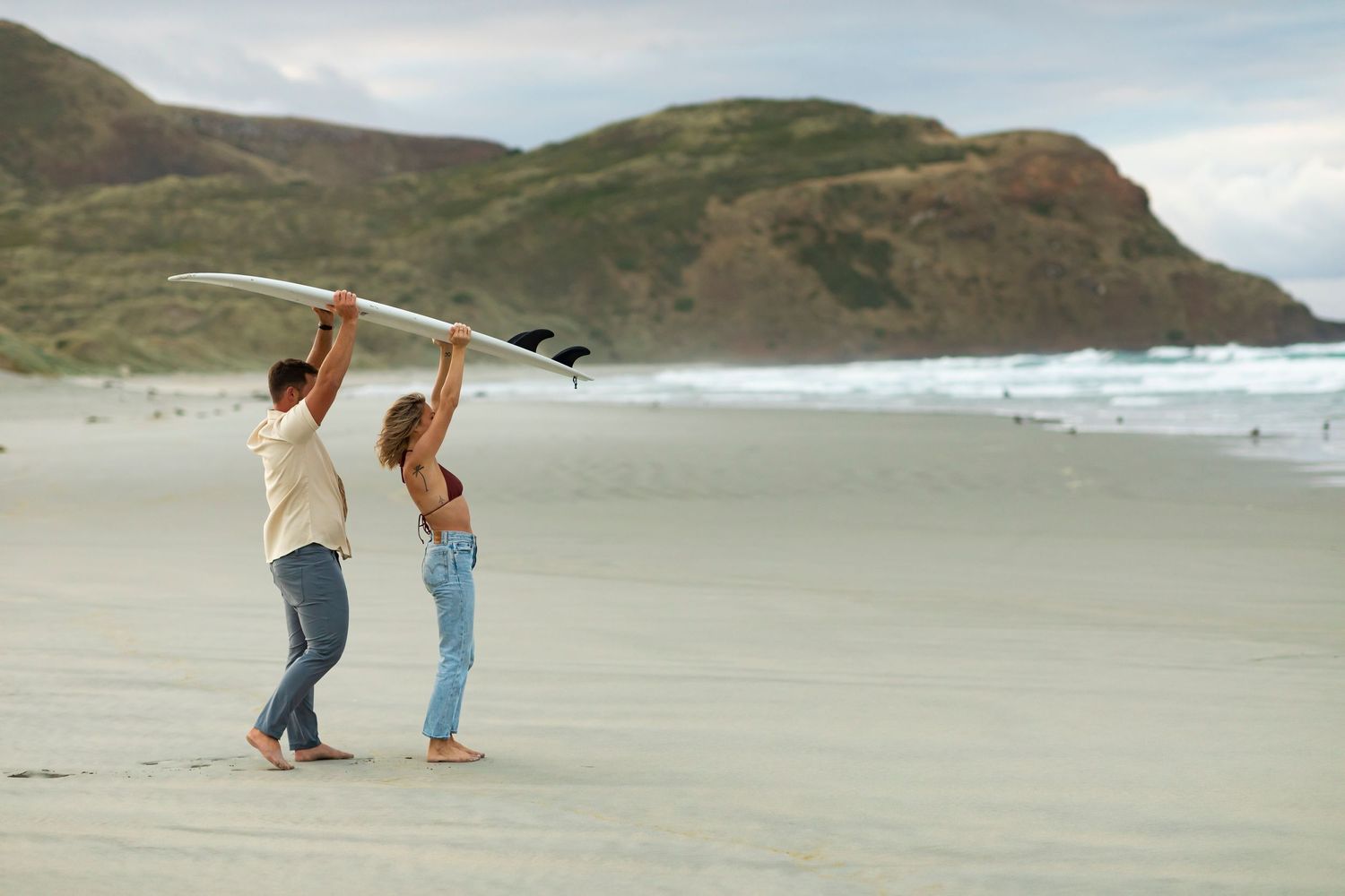 destination photoshoot of beach engagement new zealand couple carries surfboard