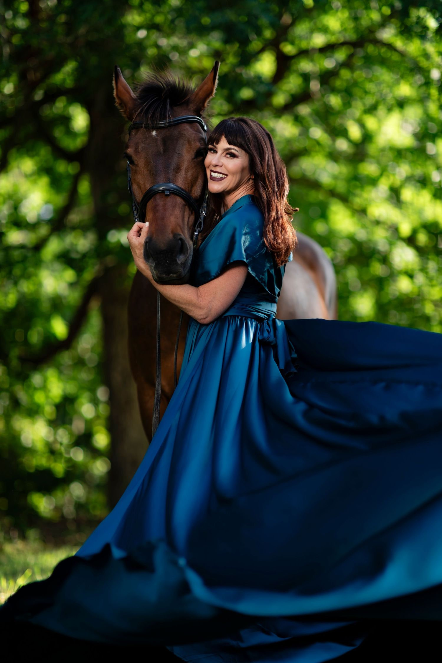 A joyful moment captured with a brown horse and flowing blue dress against lush green foliage.