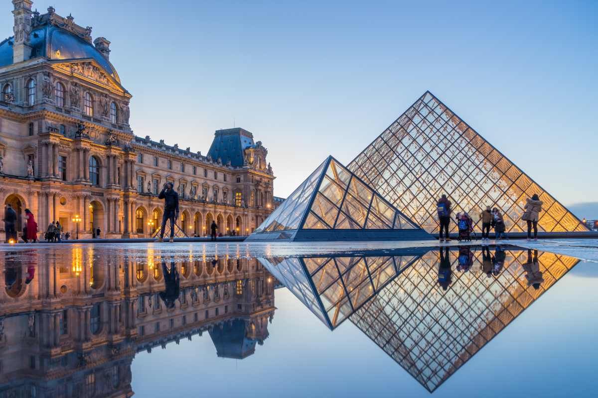 The iconic Louvre Museum pyramid reflects in a puddle during blue hour, with the historic palace facade in Paris, France.