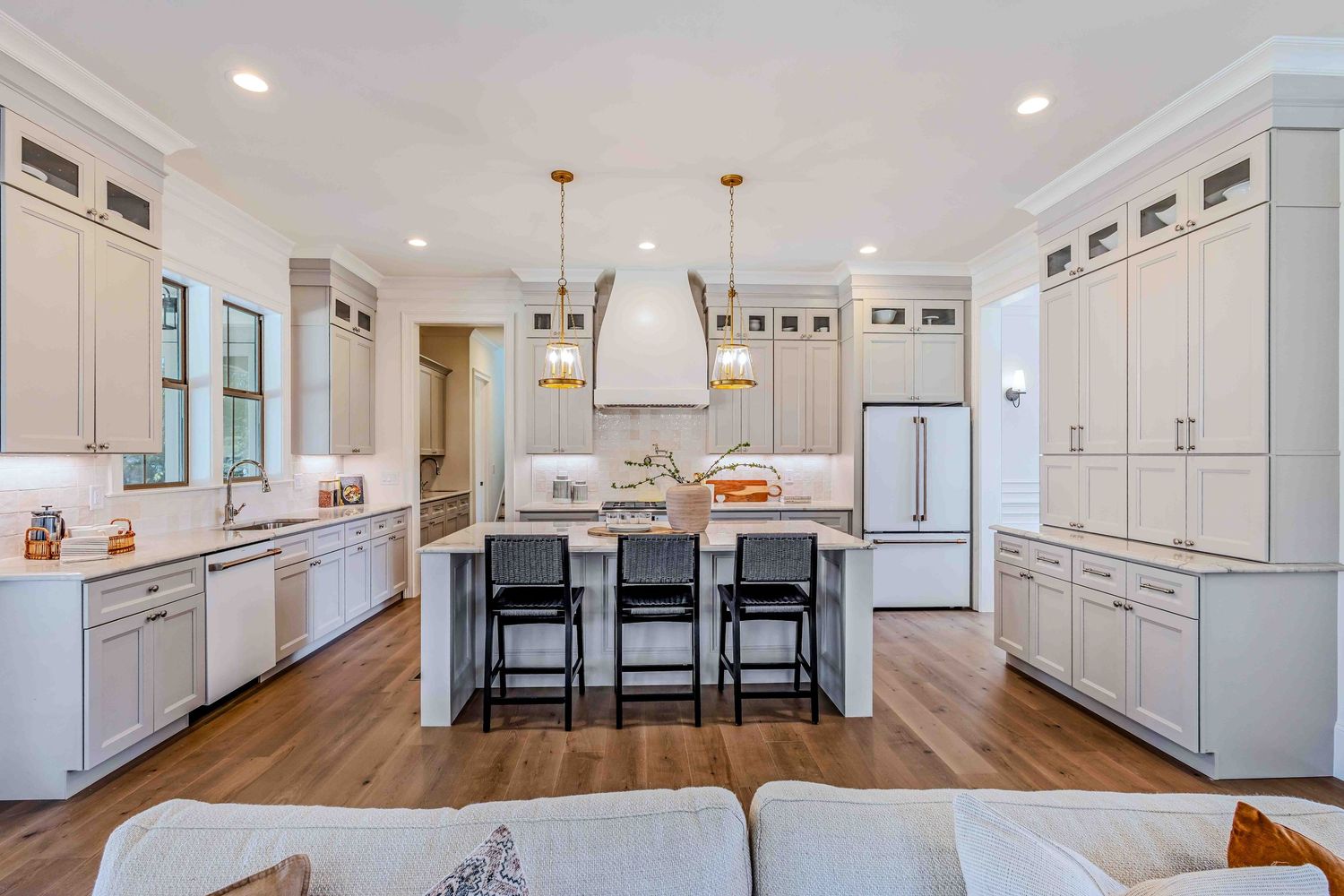 Modern white kitchen with hardwood floors, pendant lights, and black barstools at a large center island.