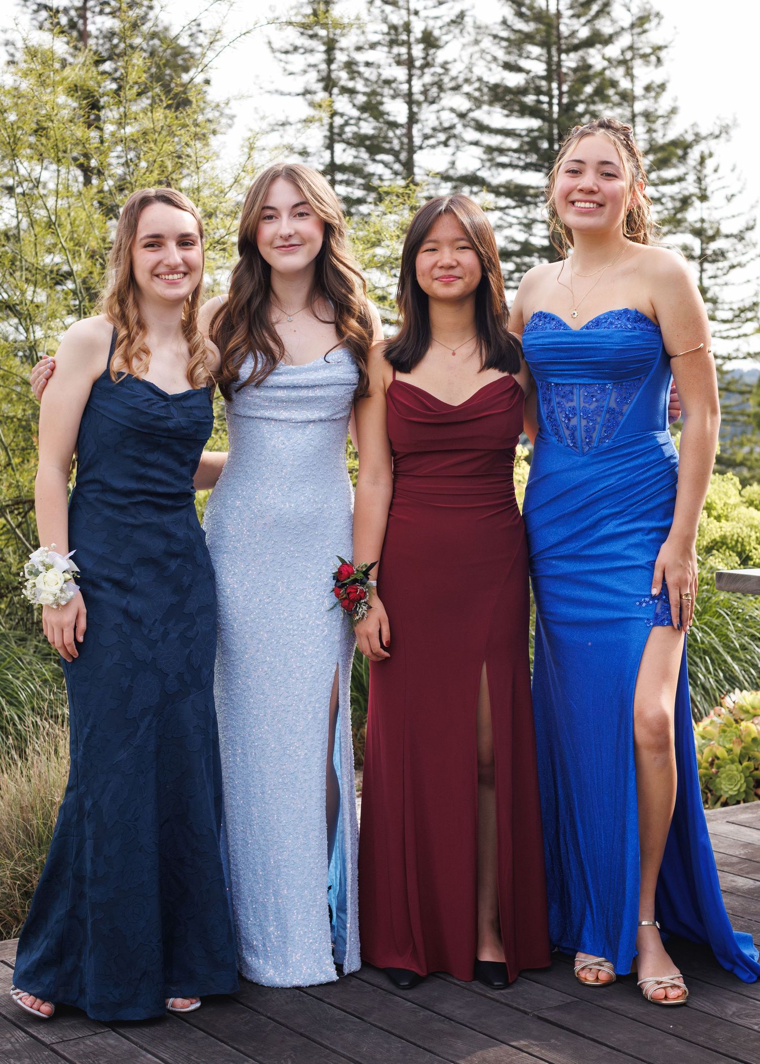 Four young women in elegant formal gowns pose together outdoors, wearing corsages at what appears to be prom.