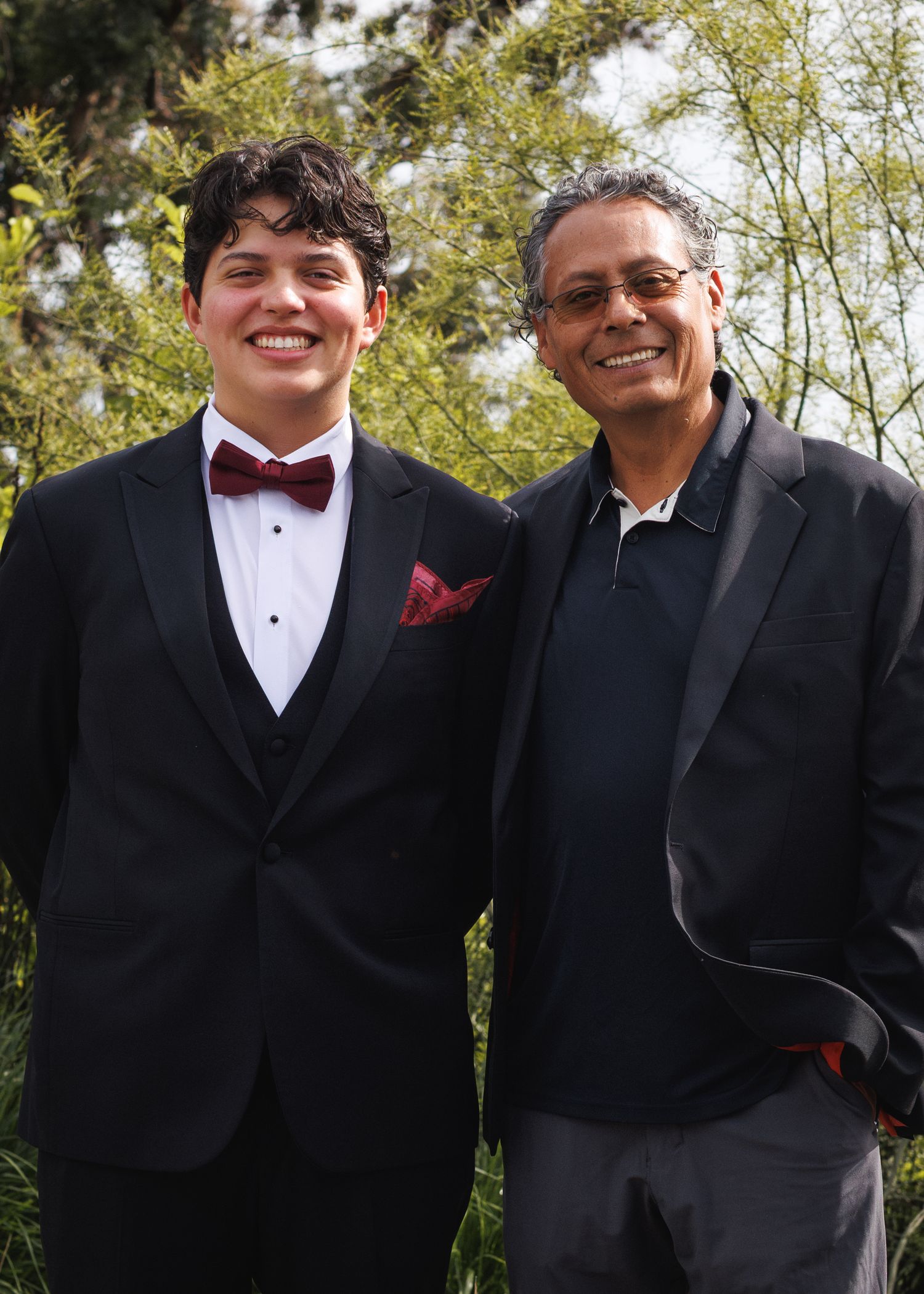 A young man in a tuxedo with bow tie poses with an older man in a navy jacket outdoors among green trees.