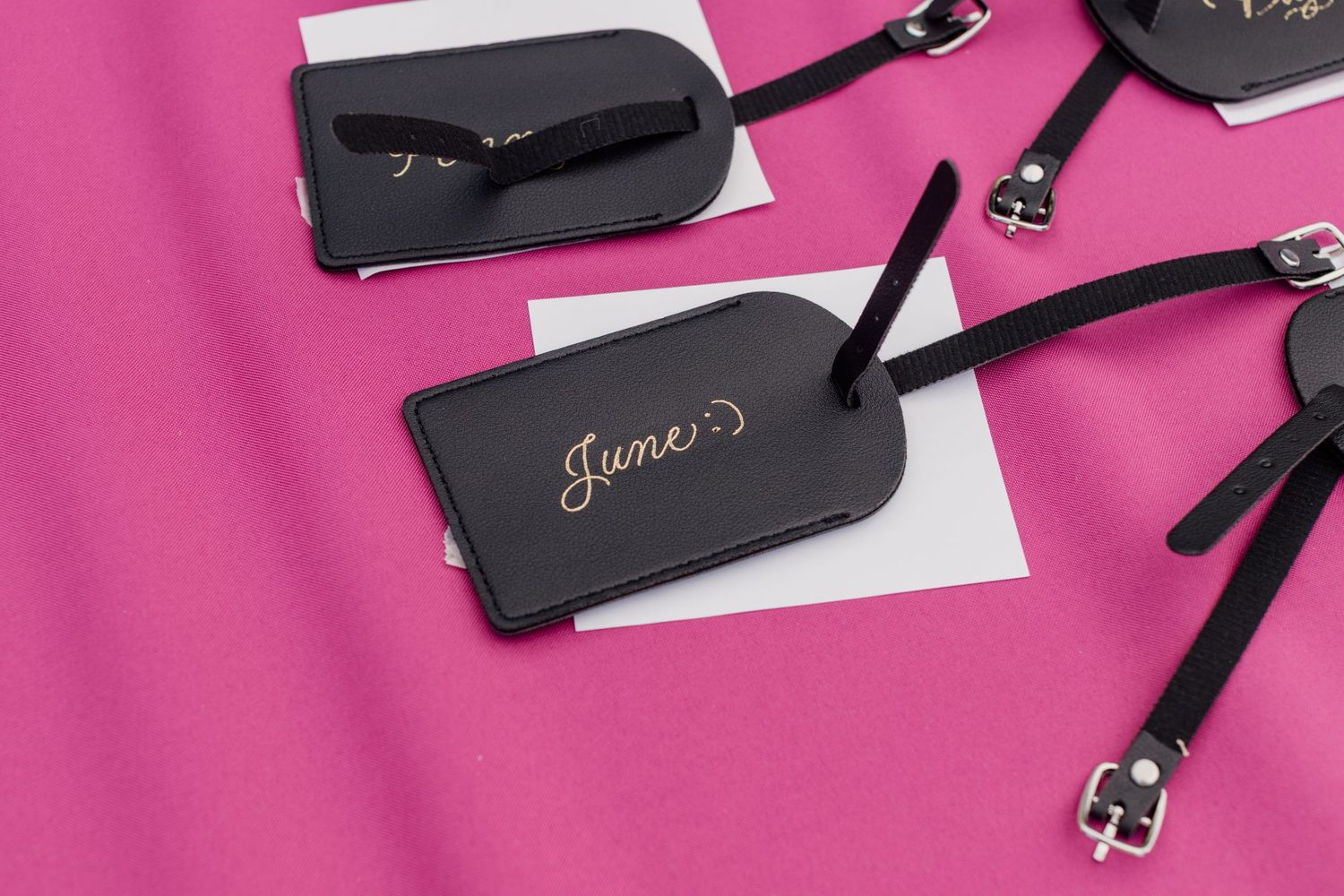 Black leather luggage tags with gold script names on a vibrant pink background, perfect for personalized travel accessories.