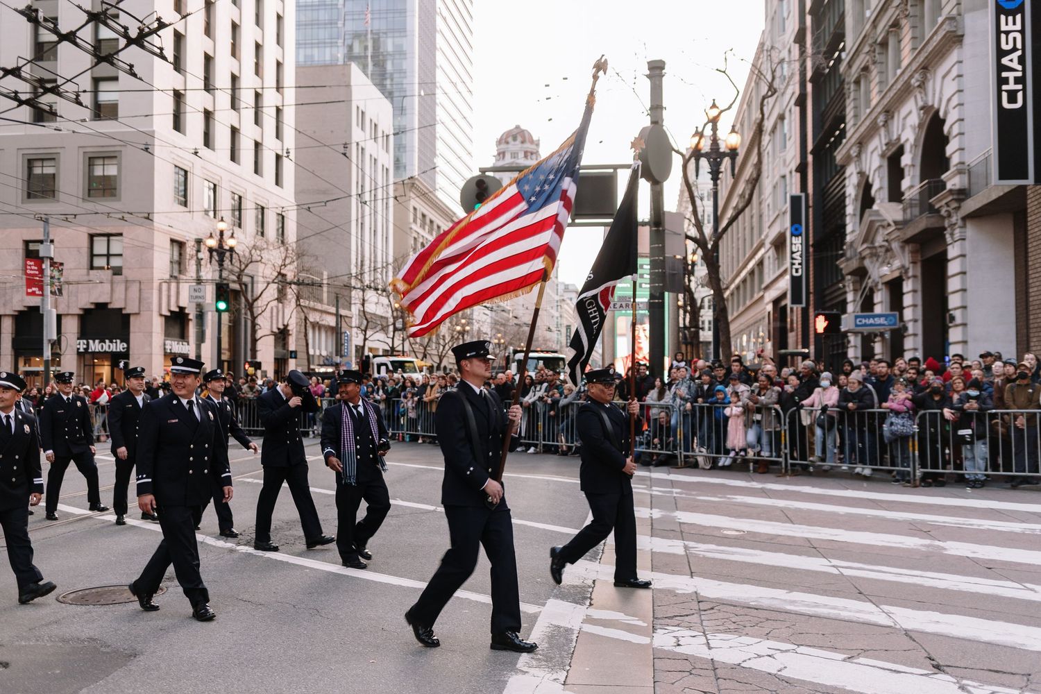 Uniformed firefighters march in a city parade carrying the American flag before a large crowd of spectators.
