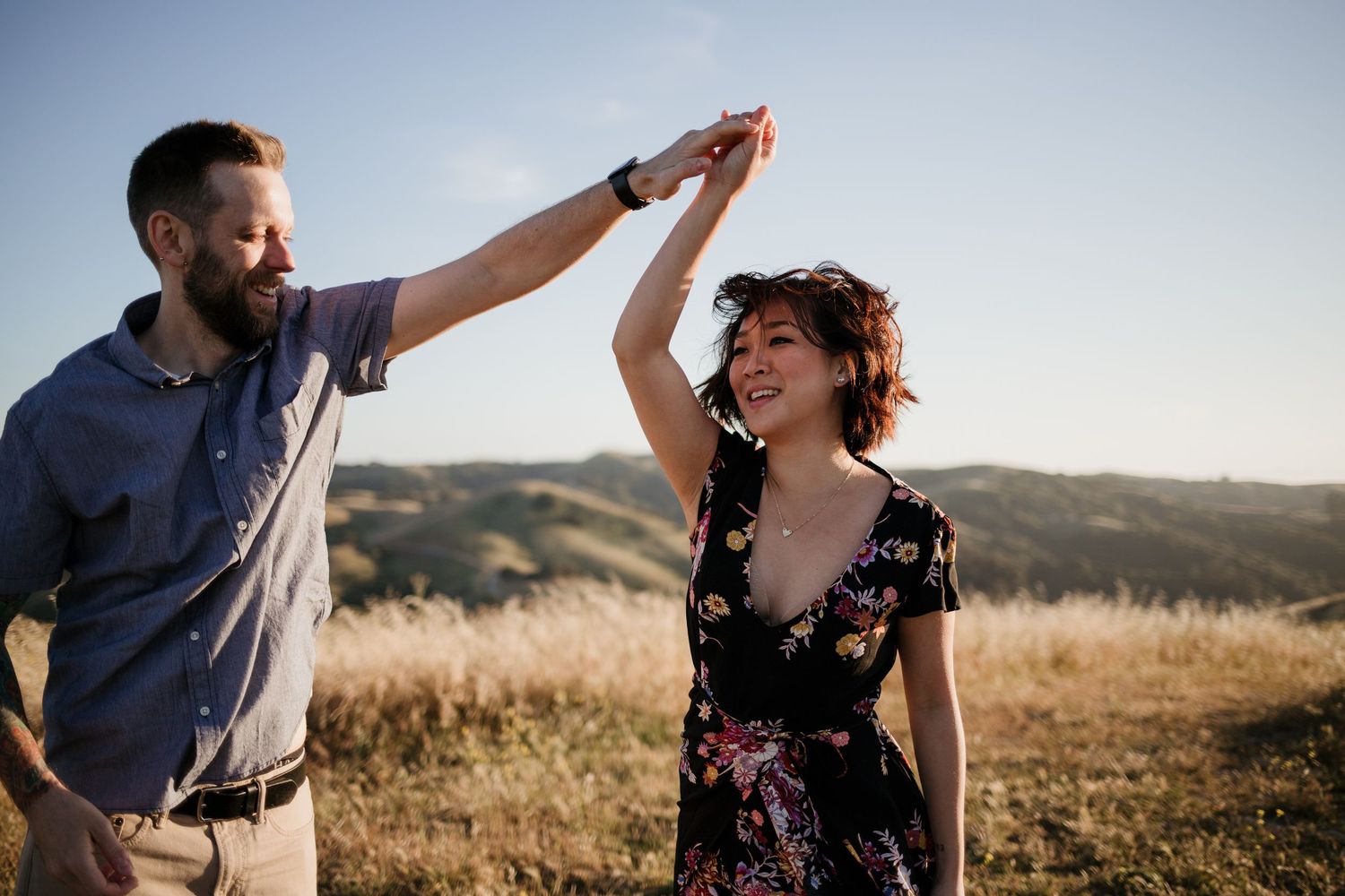 A couple dances joyfully in a sunlit field, man spinning woman in floral dress with rolling hills backdrop in Dublin, California.