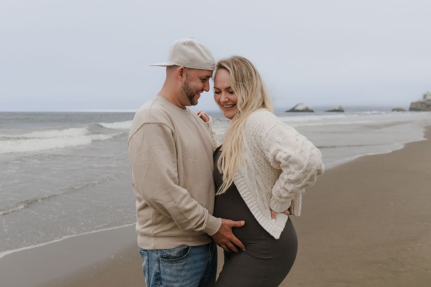 A couple in matching beige sweaters share an intimate moment on a cloudy beach.