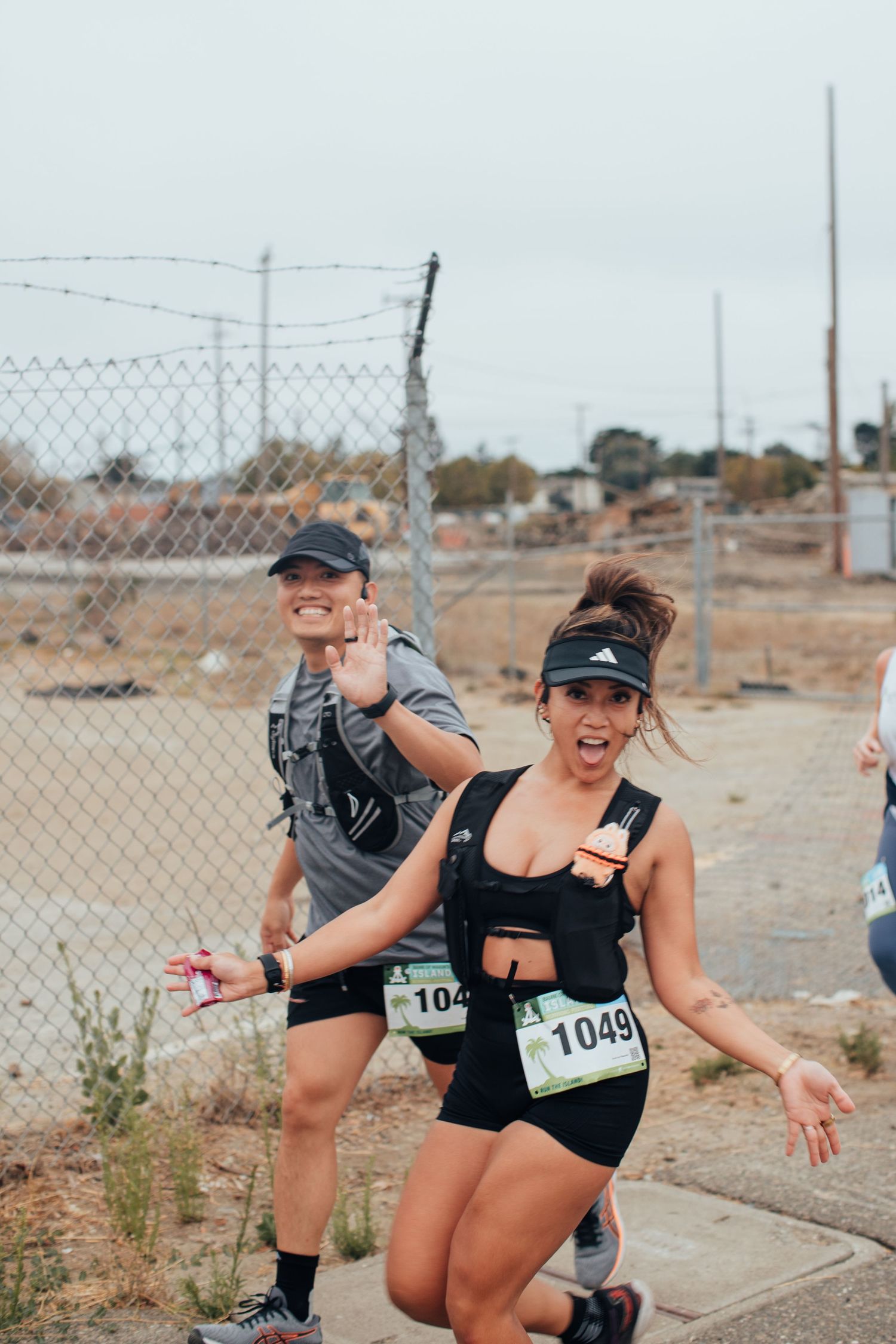 Two women wearing race bibs run in a marathon, smiling and waving at the camera near a chain-link fence.